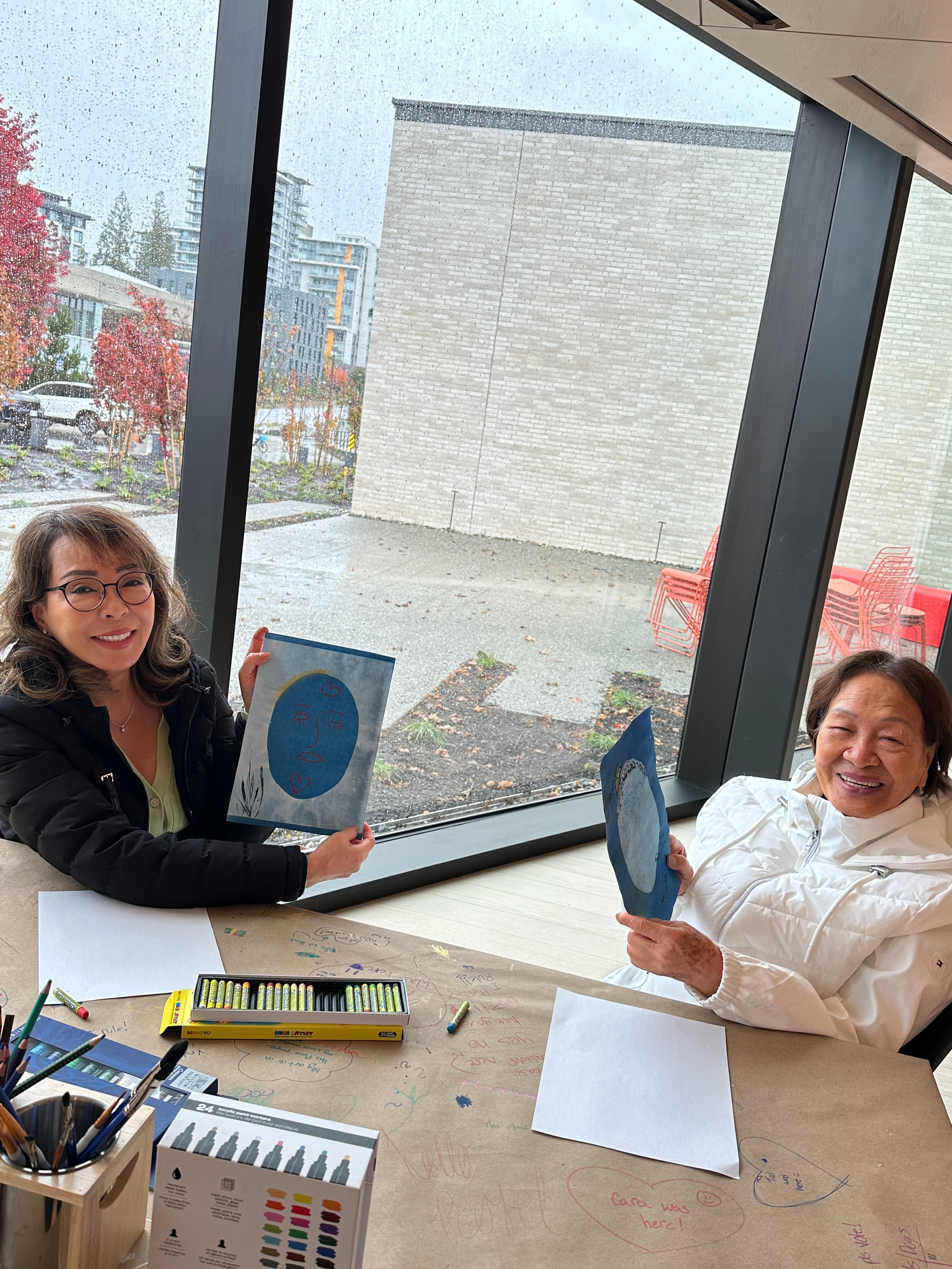 Two women are sitting at a table inside a brightly lit room with large windows, showing a rainy scene outside. They are holding up blue pieces of paper with artistic designs while smiling at the camera. The table is covered with brown paper and features scattered markers and crayons.