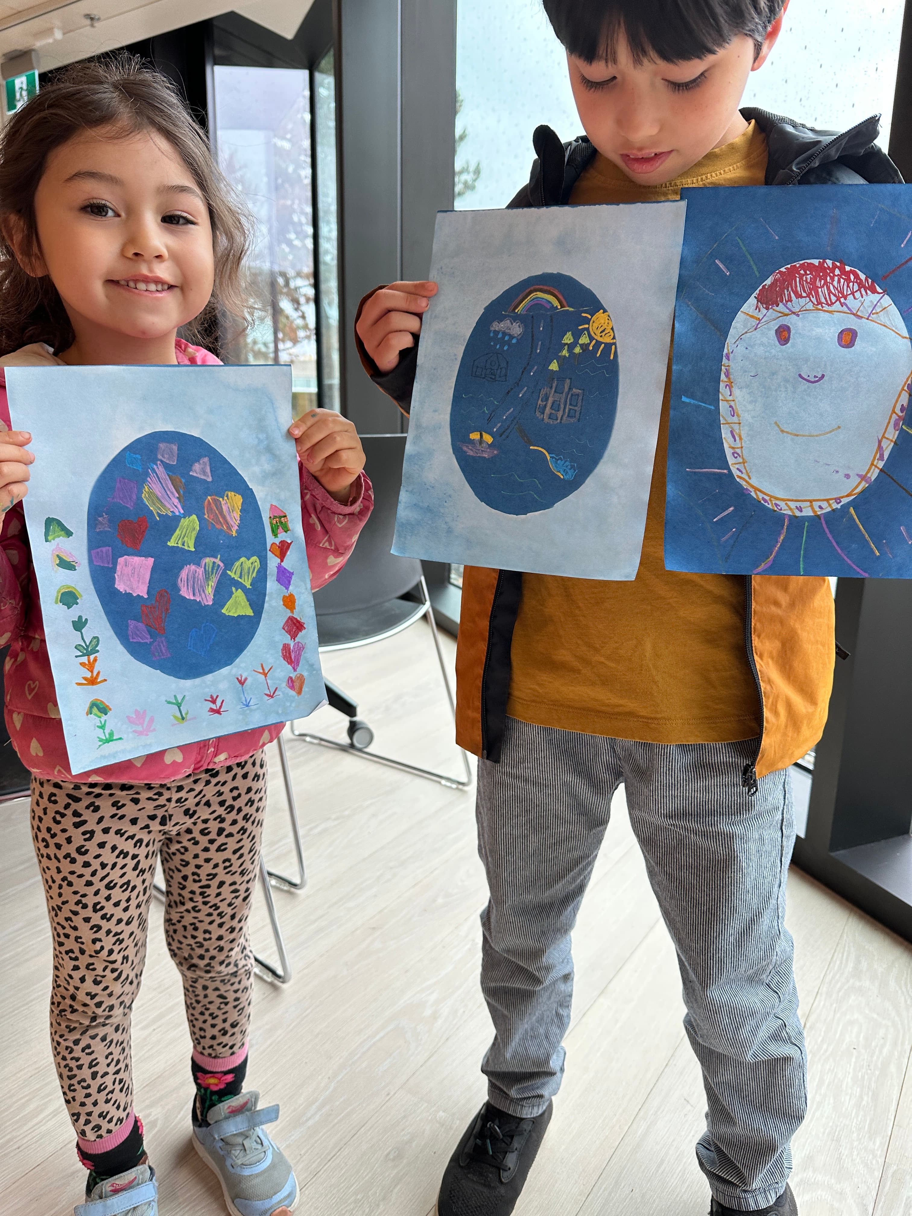 Two children are standing indoors, smiling while holding up their artwork. The girl on the left is wearing a pink jacket with heart patterns and black leopard-print leggings, while the boy on the right is dressed in a tan shirt and an orange jacket with gray pants. They are each holding a colorful drawing; the girl has a blue background with various shapes, and the boy holds two drawings: one depicting a rainbow and sun, and the other featuring a smiling face. Natural light comes through large windows in the background.