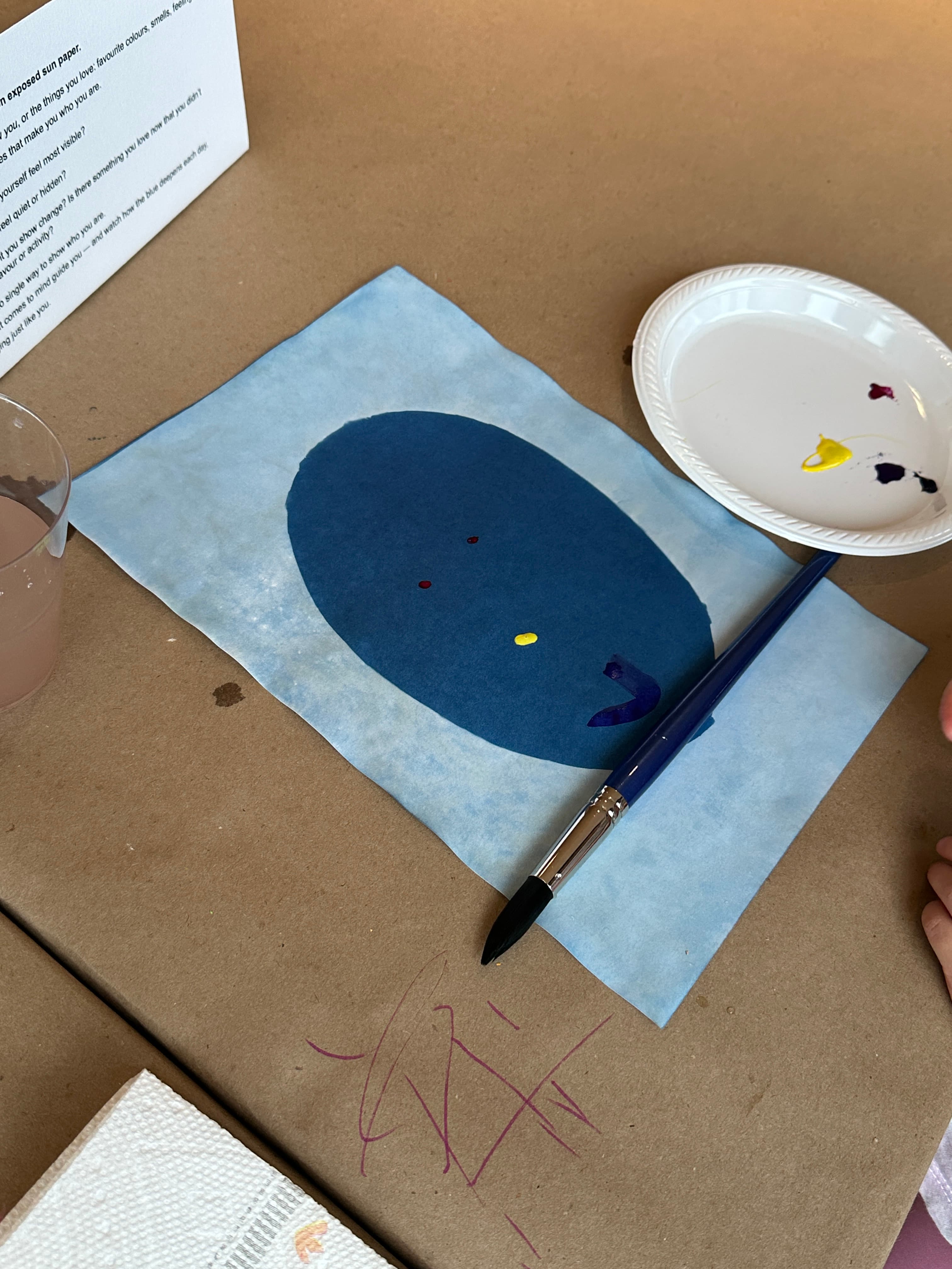A piece of blue art paper featuring an oval shape painted in a darker blue, with small dots in red and yellow. A paintbrush rests nearby, and there is a white plate with splashes of color. A clear cup containing a liquid sits in the background, and the surface is covered with brown paper showing some additional marks. A printed sheet of paper with text is partially visible in the background.