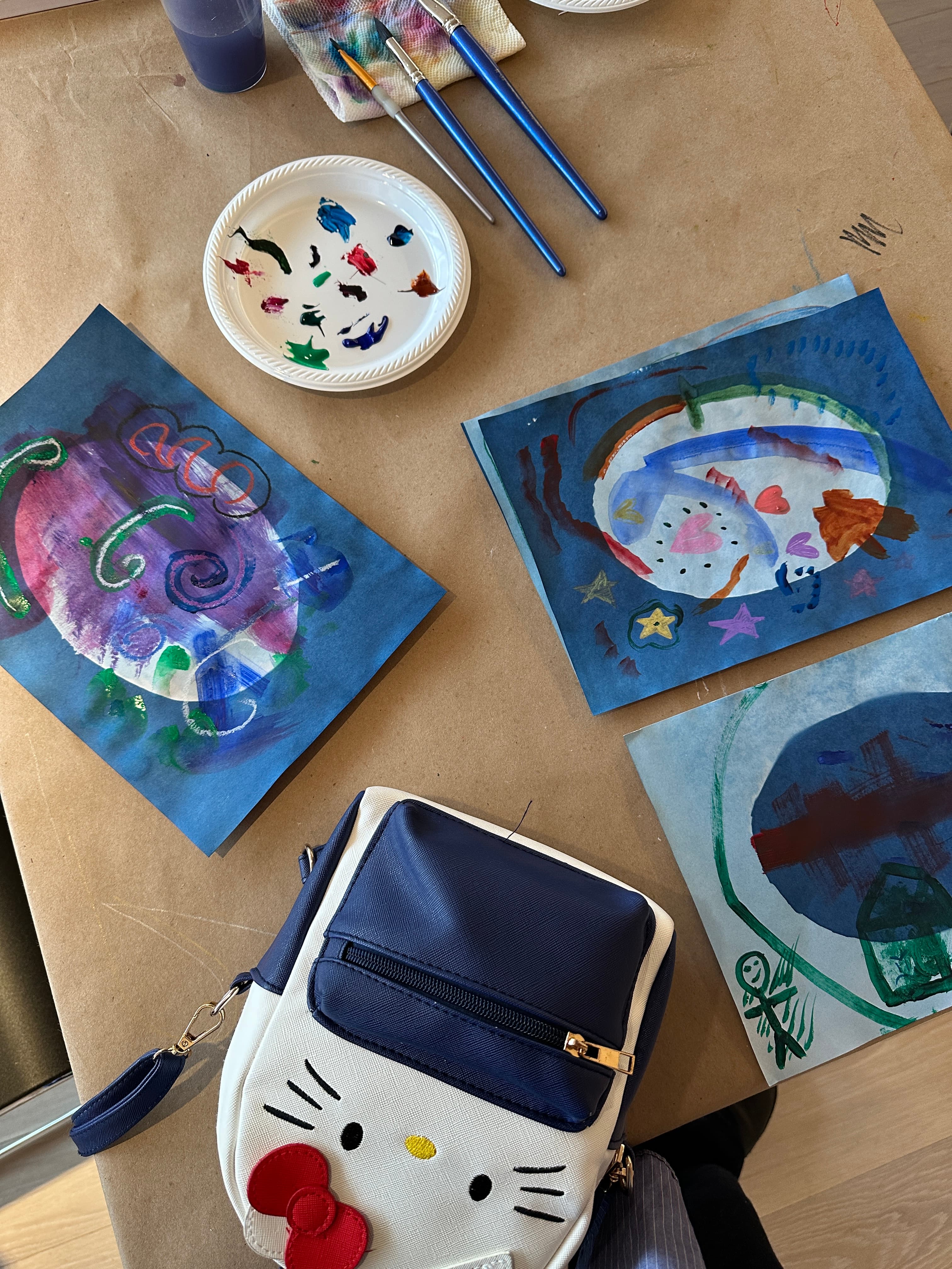 A tabletop covered in brown paper features three colorful paintings on blue paper, depicting abstract designs and a winter scene. A white plate holds various paint colors, and several paintbrushes are nearby, along with a small purple container and a cloth. A Hello Kitty-themed bag is visible in the lower corner of the image.