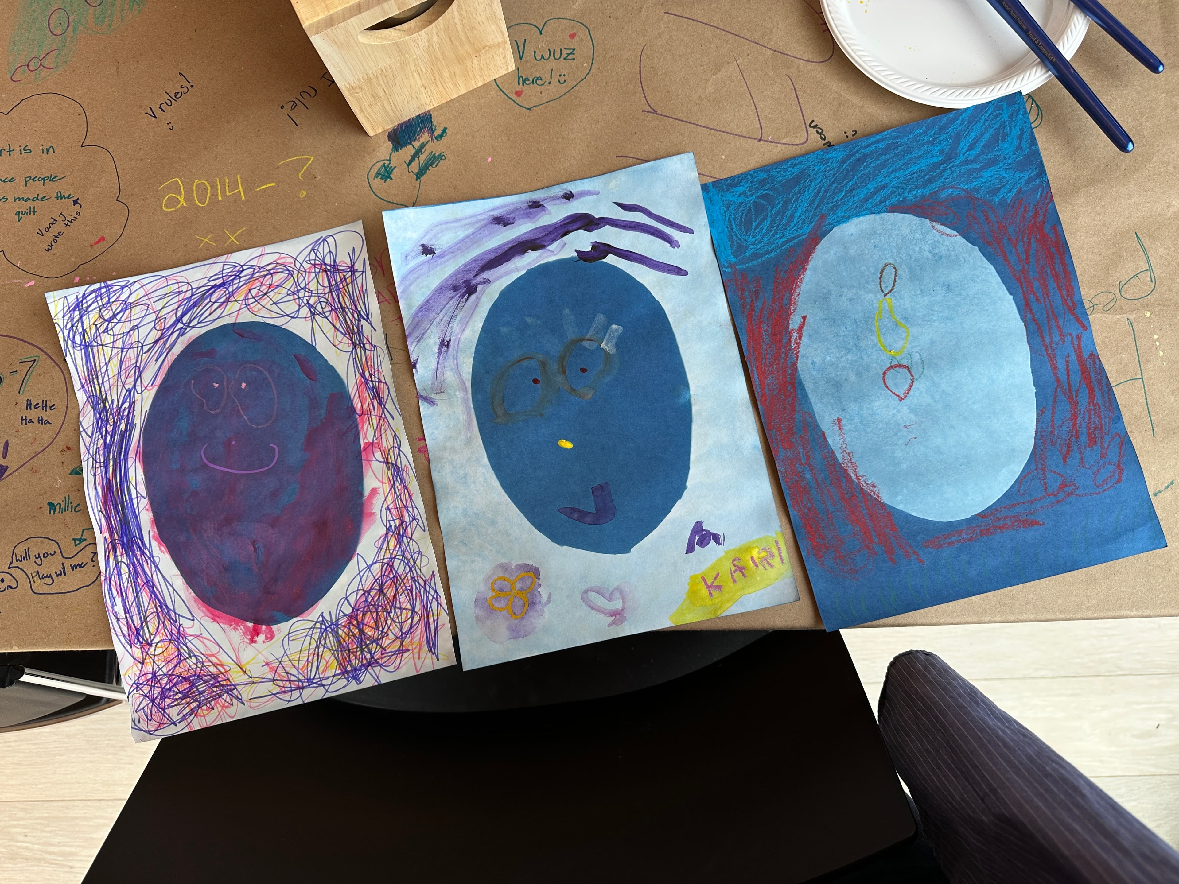 Three colorful drawings are displayed on a brown table covered with doodles and text. The first drawing on the left features a large purple oval with facial features drawn in a simple style. The middle drawing has a blue background with a smiling face and glasses, along with a small flower. The drawing on the right depicts an oval shape against a blue and red background, with colorful shapes above it. Nearby, there are a couple of paintbrushes and a paper plate.