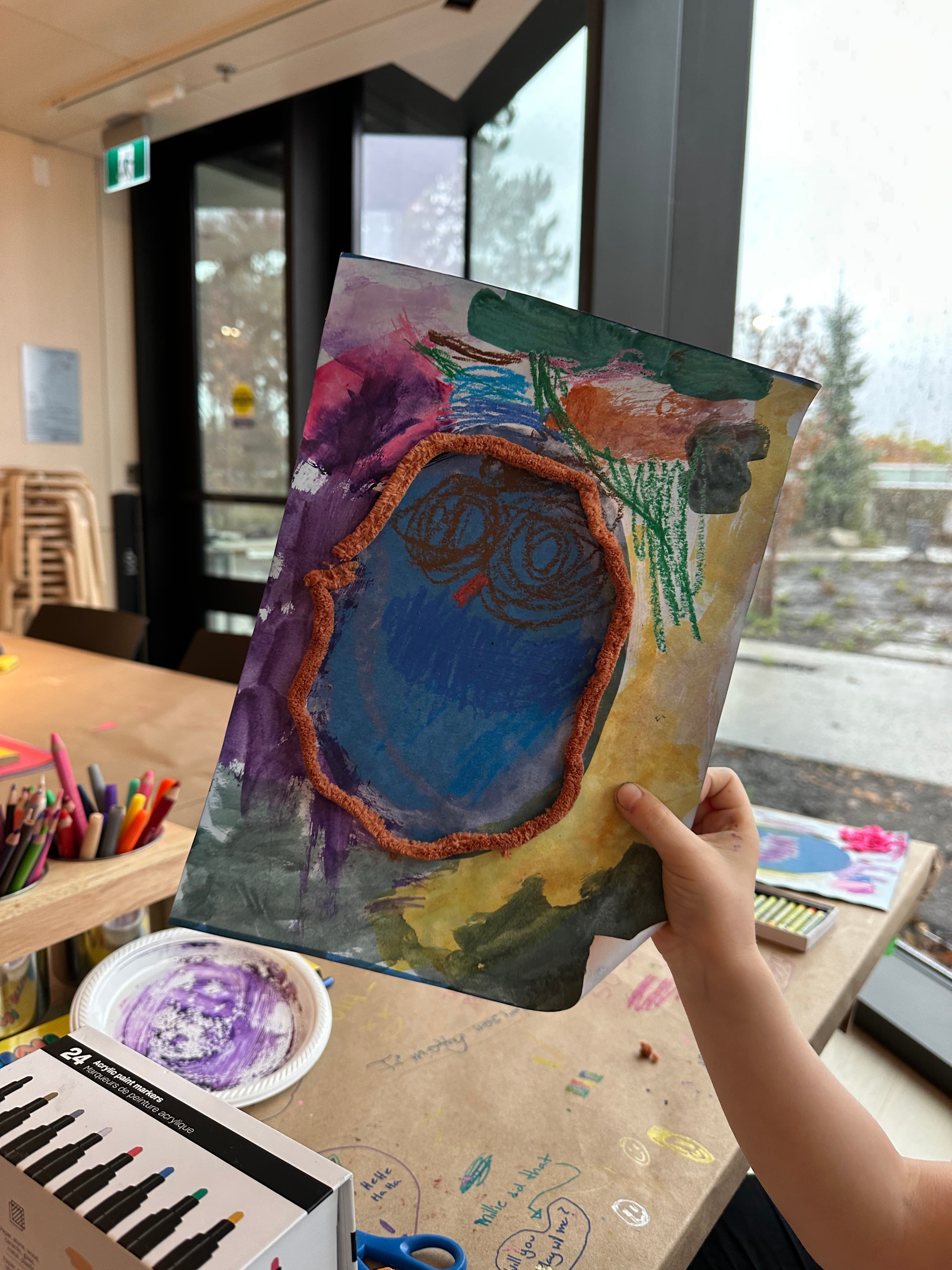 A child is holding a colorful, abstract artwork that depicts a blue face surrounded by splashes of different colors, including purple, green, and orange. The background is textured with brush strokes, and there are art supplies on a table in the foreground, including markers and plates for mixing. The scene is in a well-lit room with large windows showing rainy weather outside.