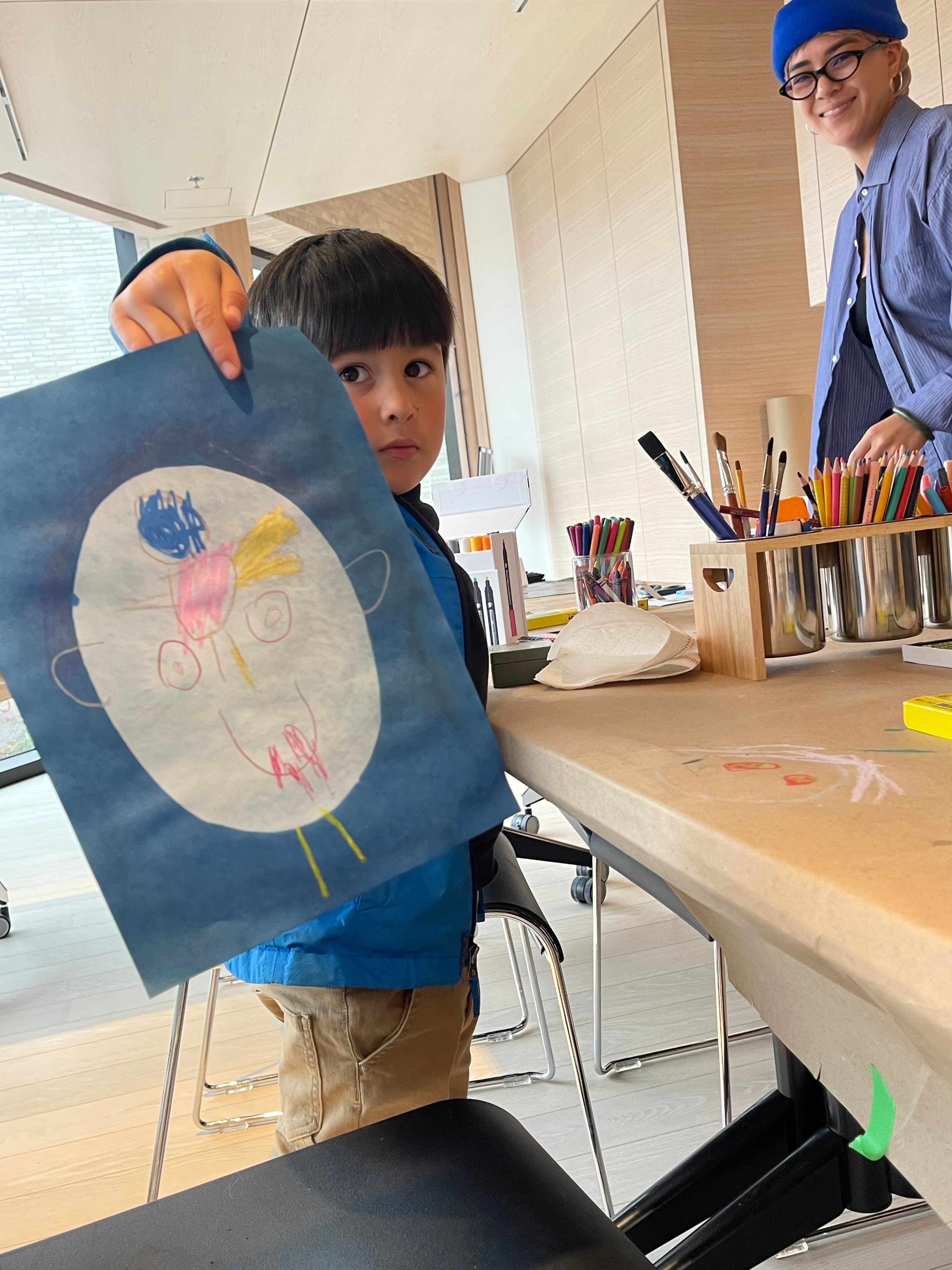 A young boy with short dark hair is holding a colorful drawing on blue paper, depicting a face with various facial features. He is wearing a blue jacket and looks towards the camera with a serious expression. In the background, an adult woman with short blonde hair, wearing a blue beanie and glasses, smiles while standing near a table filled with art supplies, including markers and brushes. The setting has large windows and a light wooden interior.