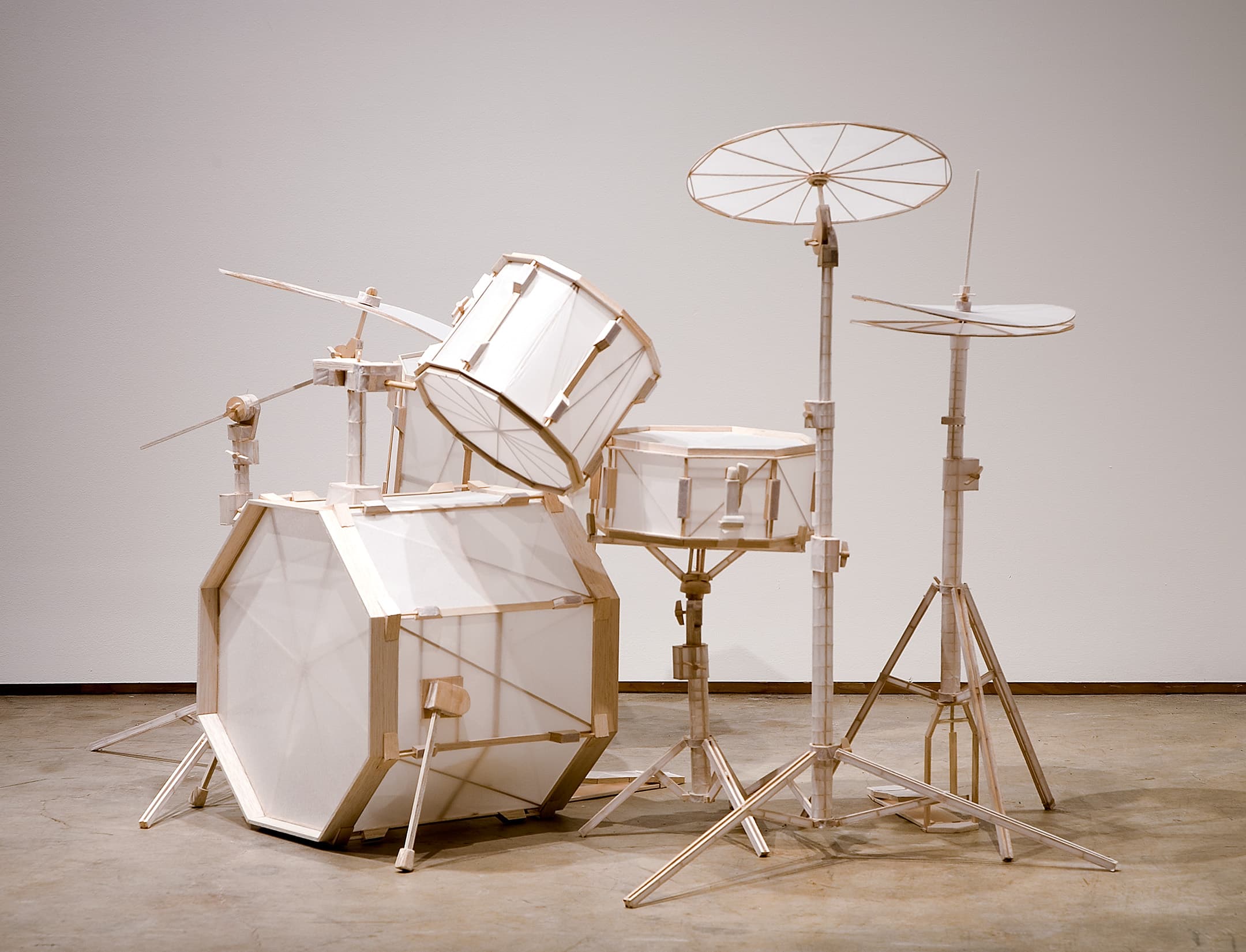 A sculpture of a drum set made from white paper and wooden elements. The kit includes a bass drum, snare drum, two cymbals, and a tom-tom, all mounted on stands. The drums have a geometric design with hexagon and circular shapes, and the overall structure appears lightweight and artistic. The background is a plain white wall, contrasting with the sculptural piece.
