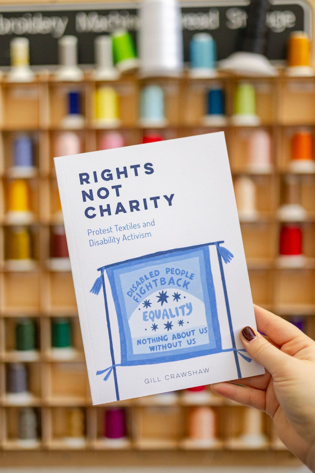 A hand is holding a book titled "Rights Not Charity: Protest Textiles and Disability Activism" by Gill Crawshaw. The cover features a blue illustration of a banner with the text: "Disabled People Fight Back Equality Nothing About Us Without Us." In the background, there is a display of colorful spools of thread arranged on a wooden shelf.