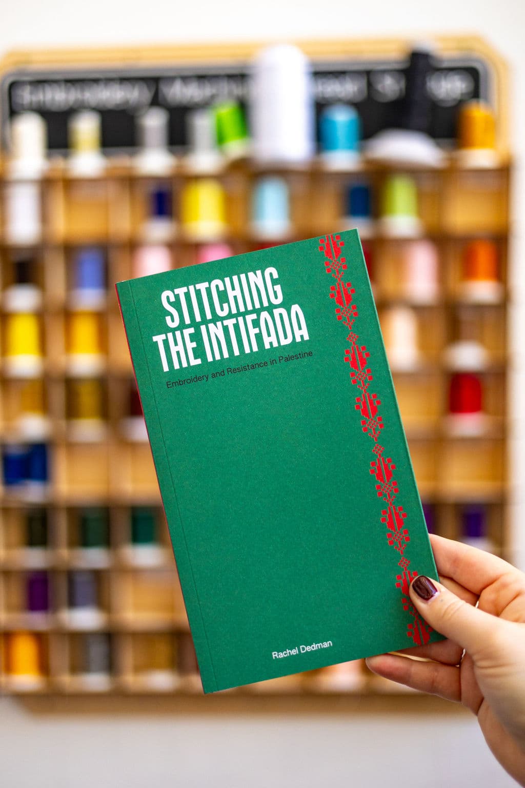 A hand holds a green book titled "Stitching the Intifada: Embroidery and Resistance in Palestine," with a decorative red pattern along the spine. In the background, a wooden shelf displays various spools of colorful thread.