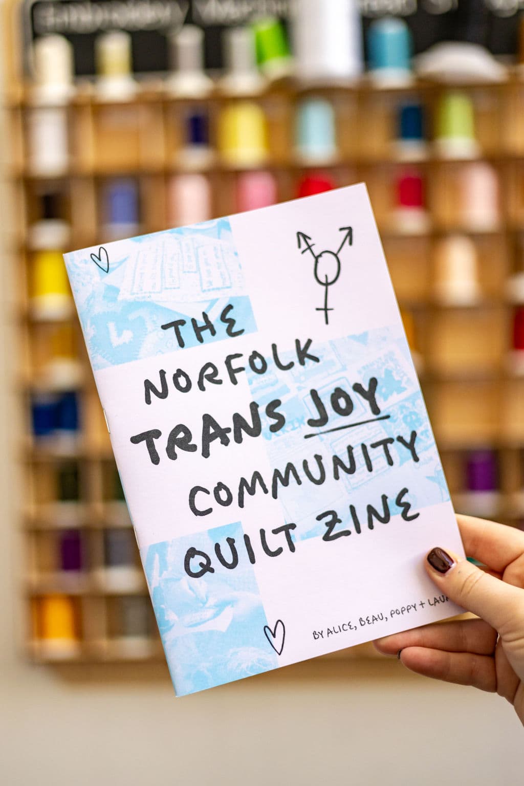 A person holds a zine titled "The Norfolk Trans Joy Community Quilt Zine." The cover features a pastel background with hand-drawn text in black, and a transgender symbol. In the background, a wooden board displays various colorful sewing threads.