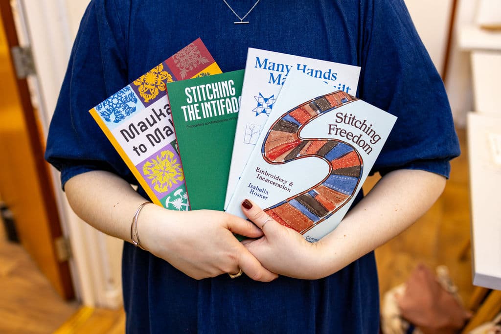 A person holds four colorful books against their chest. The titles visible include "Mauka to Makai," "Stitching the Intifada," "Many Hands Make Light Work," and "Stitching Freedom." The person is wearing a blue garment, and the background features a wooden floor and blurred furniture.