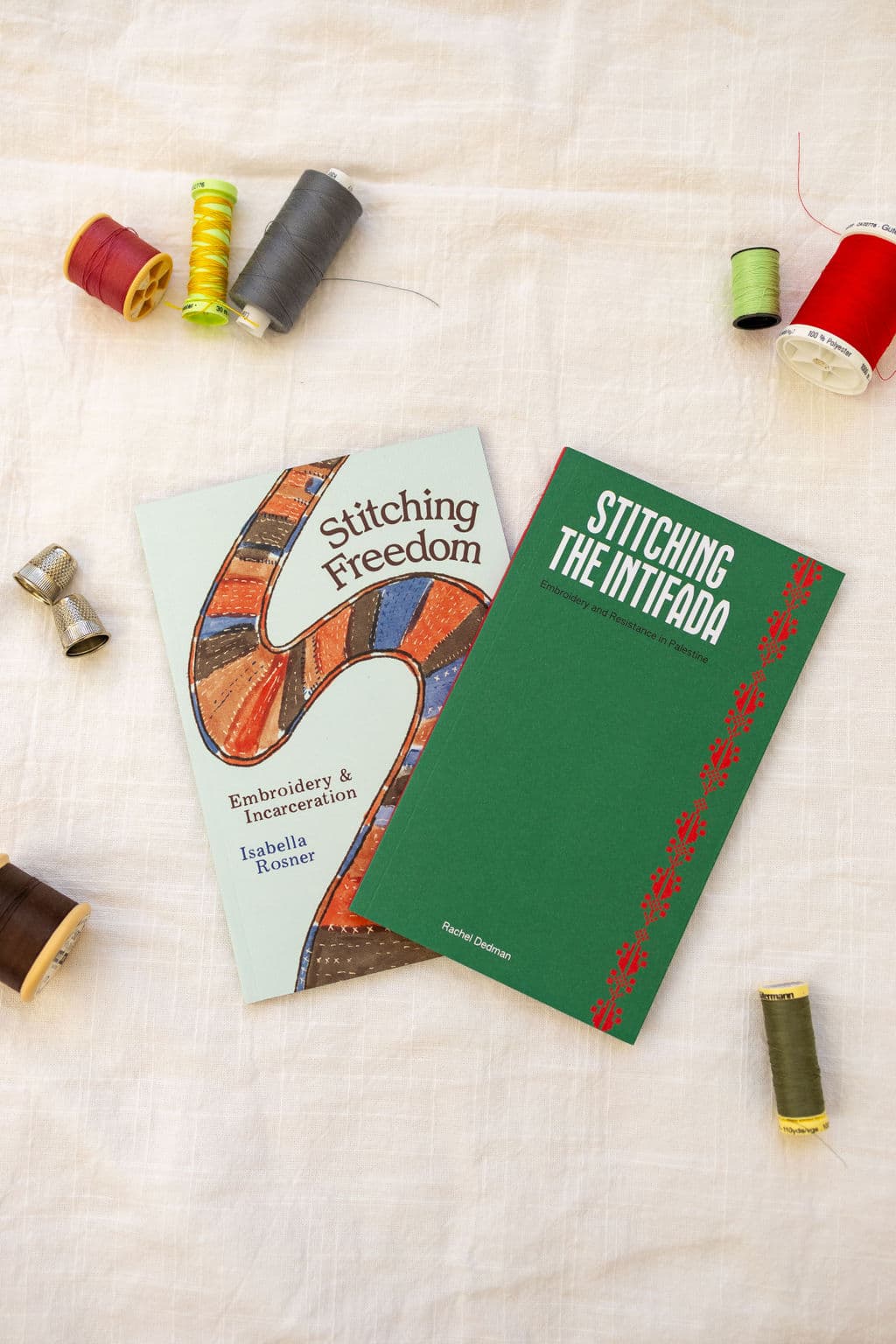 A flat lay image featuring two books: "Stitching Freedom" by Isabella Rosner with a colorful embroidered design on the cover, and "Stitching the Intifada" by Rachel Dedman with a green cover and red decorative elements. Surrounding the books are several spools of thread in various colors, including red, yellow, green, and gray, along with a thimble and a wooden spool. The background is a light textured fabric.