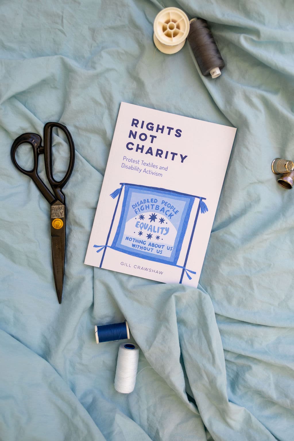 A pair of black scissors lies next to a book titled "Rights Not Charity" by Gill Crawshaw, which discusses protest textiles and disability activism. The book is open to a page featuring a blue illustration with the message "Disabled People Fight Back Equality: Nothing About Us Without Us." Surrounding the book are spools of thread in blue and white on a light blue fabric backdrop.