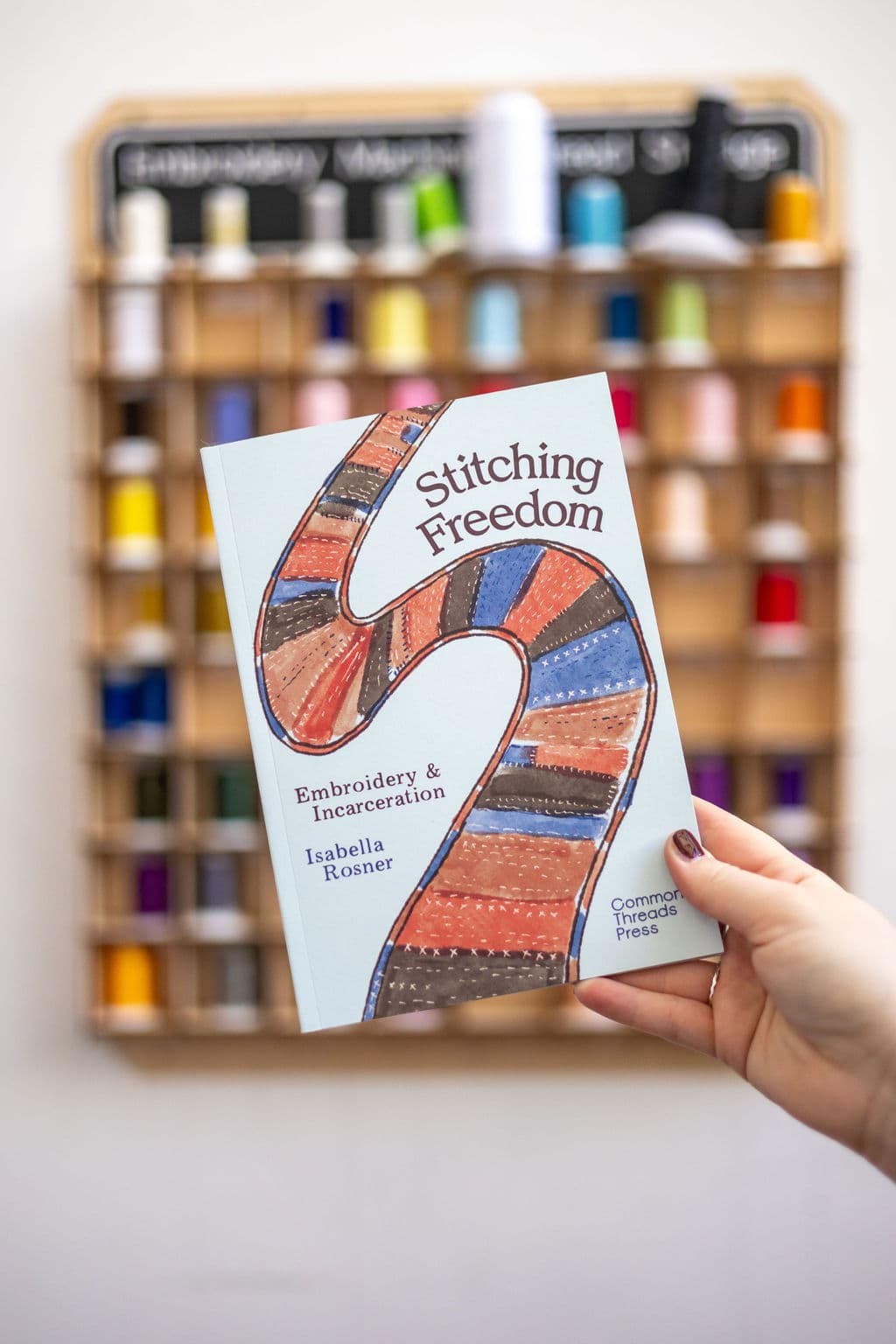 A hand holding the book titled "Stitching Freedom: Embroidery & Incarceration" by Isabella Rosner. The cover features a colorful, stitched ribbon design against a light blue background. In the background, a wooden shelf displays various spools of thread in different colors.