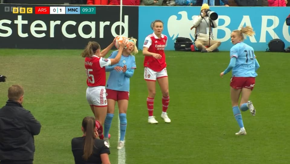 A women's football match scene shows players on the field during a game. One player in a red jersey pushes the soccer ball against the face of a player in a light blue jersey. Two teammates stand nearby. The scoreboard indicates the score is Arsenal 0, Manchester City 1, and a photographer is seen capturing the action in the background. The match is being played on a green pitch with branding from a sponsor visible.