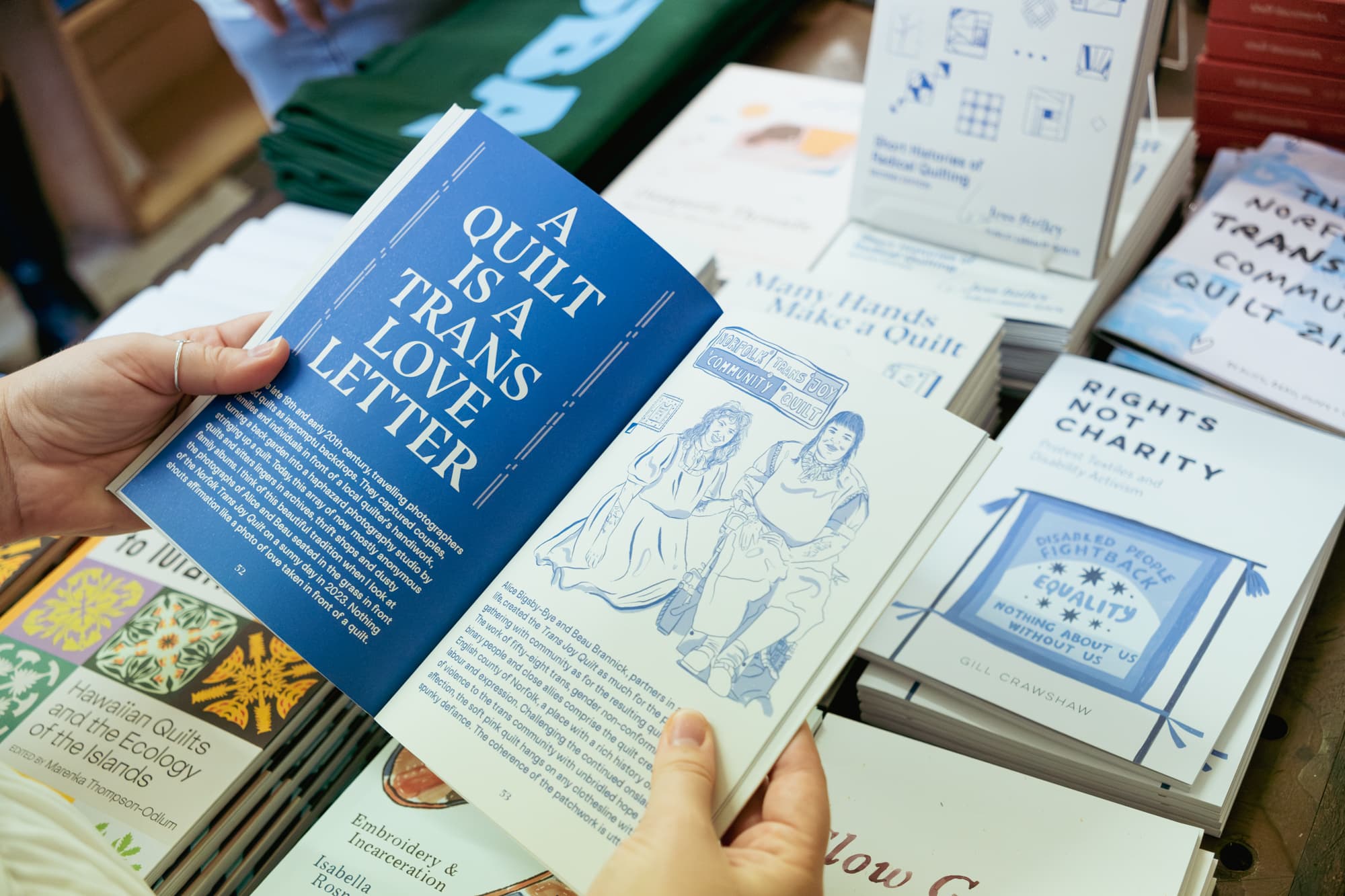 A hand holds an open book displaying a page with the title "A QUILT IS A TRANS LOVE LETTER" in bold white text against a blue background. Below, there's an illustration of two characters sitting together, along with accompanying text. The book is positioned above a table covered with various other books related to quilting, activism, and history.