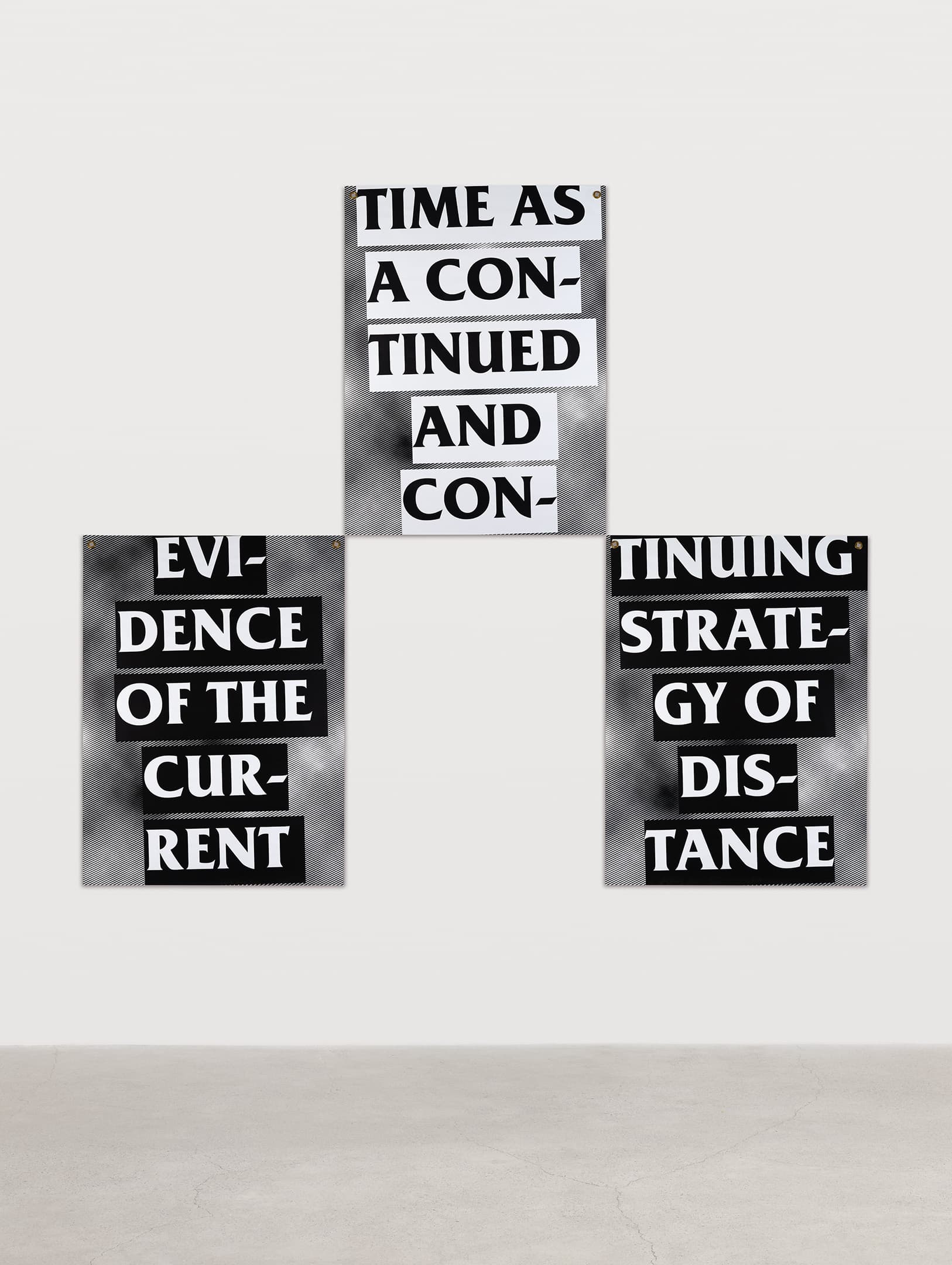 Three posters arranged on a light gray wall, featuring bold, black text on a contrasting background. The top poster reads "TIME AS A CONTINUED AND CON-," the left poster states "EVIDENCE OF THE CURRENT," and the right poster says "TINUING STRATEGY OF DISTANCE." The floor is light concrete.