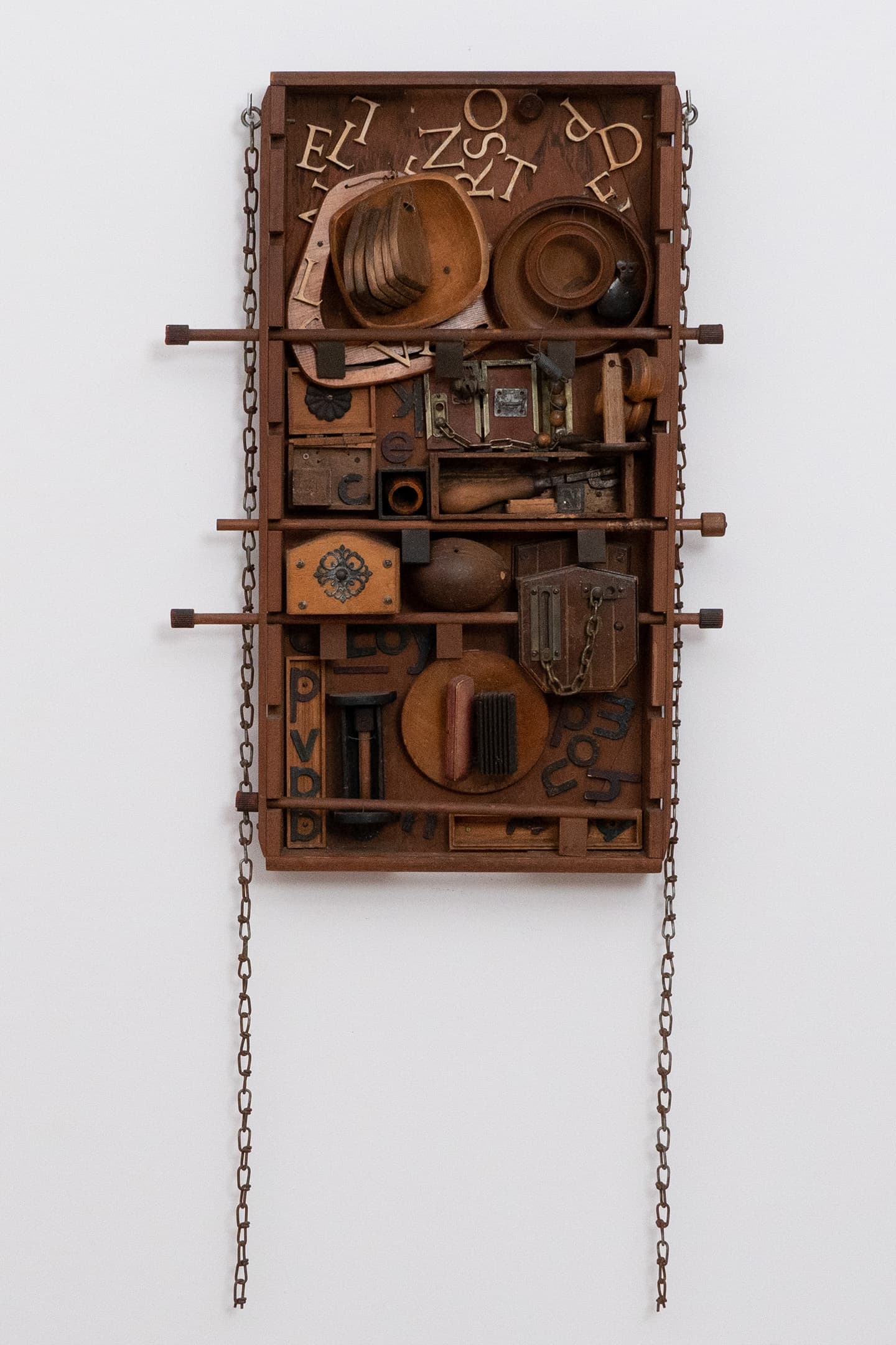 A wooden assemblage art piece consists of various objects, including hats, small boxes, letters, and other items, arranged in a grid on a rectangular board. The artwork is framed with chains on either side and features different textures and shapes, creating a dynamic visual composition.