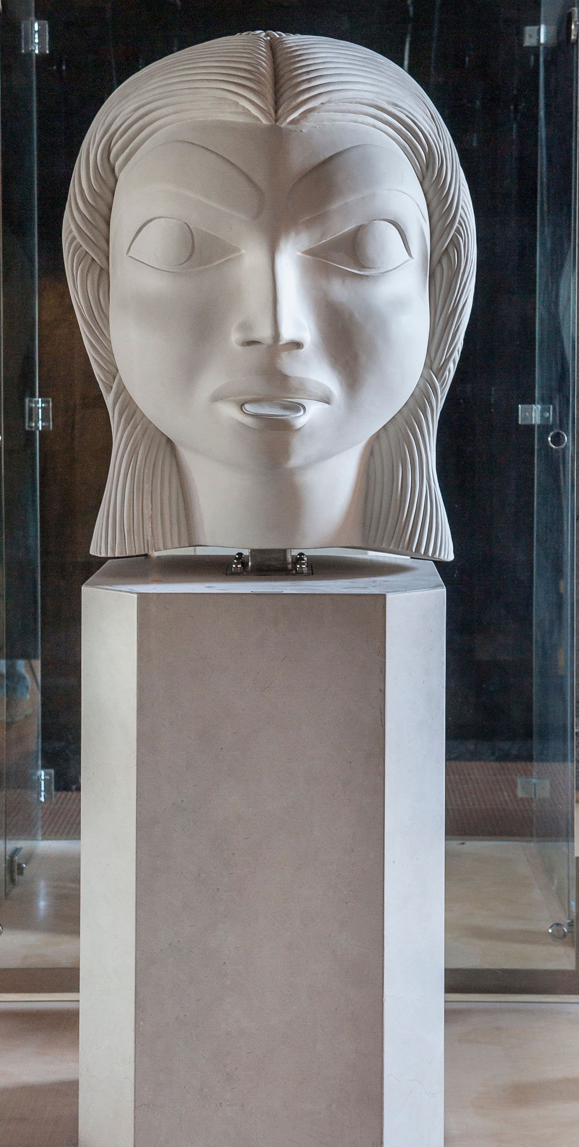 A large, sculpted head with pronounced features and textured hair sits on a tall, rectangular pedestal. The head has an expressionless face, with large, almond-shaped eyes and a small mouth. The sculpture is displayed in a glass enclosure, with a dark background that contrasts with the lighter materials of the head and pedestal.
