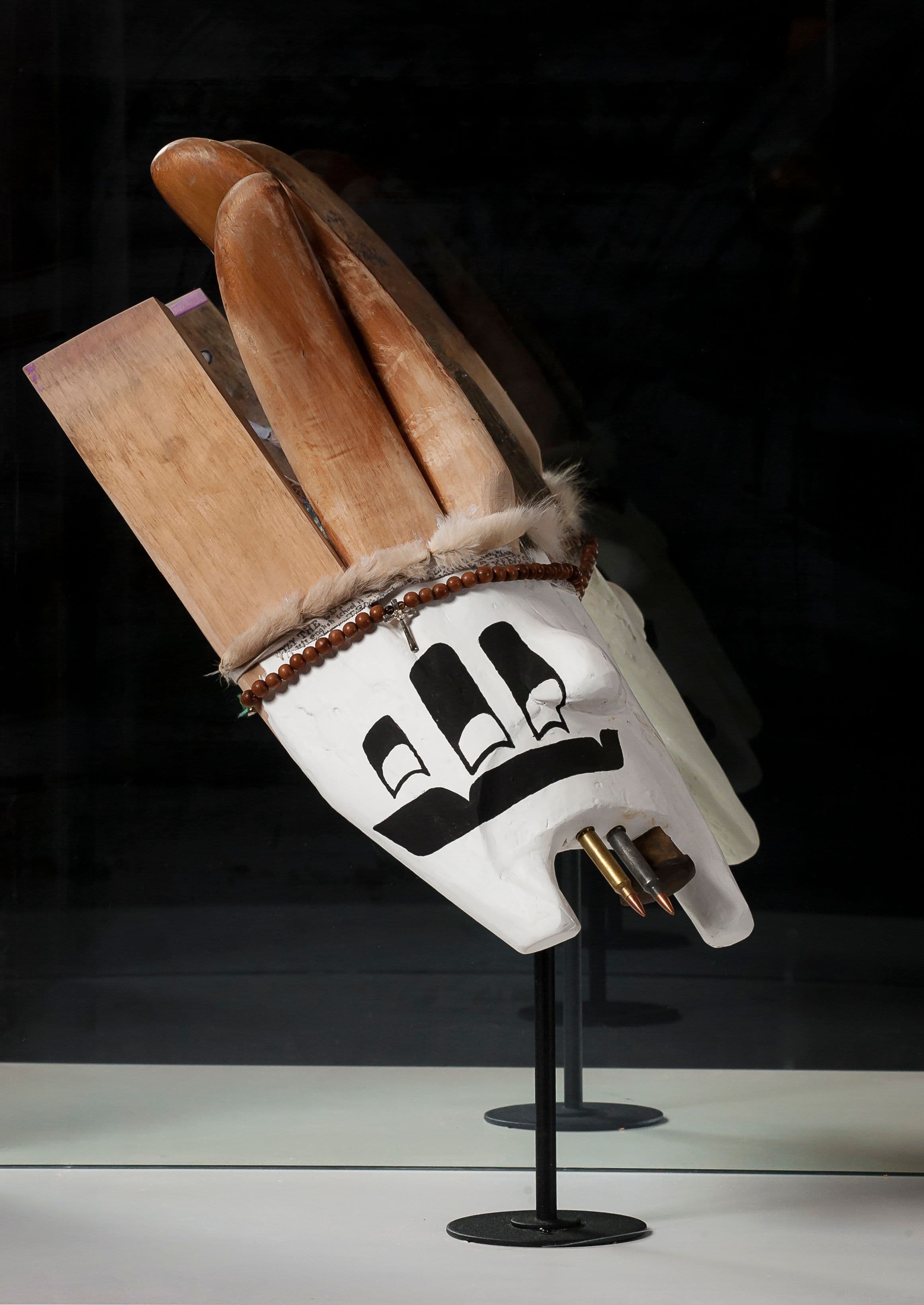A white sculpture of a mask, featuring black markings on the surface. The mask holds wooden objects resembling blades, with three rounded wooden forms extending upwards. A decorative band with fur and beads encircles the forehead area, with small metallic objects protruding from the lower half. The sculpture is displayed on a black stand against a dark background.