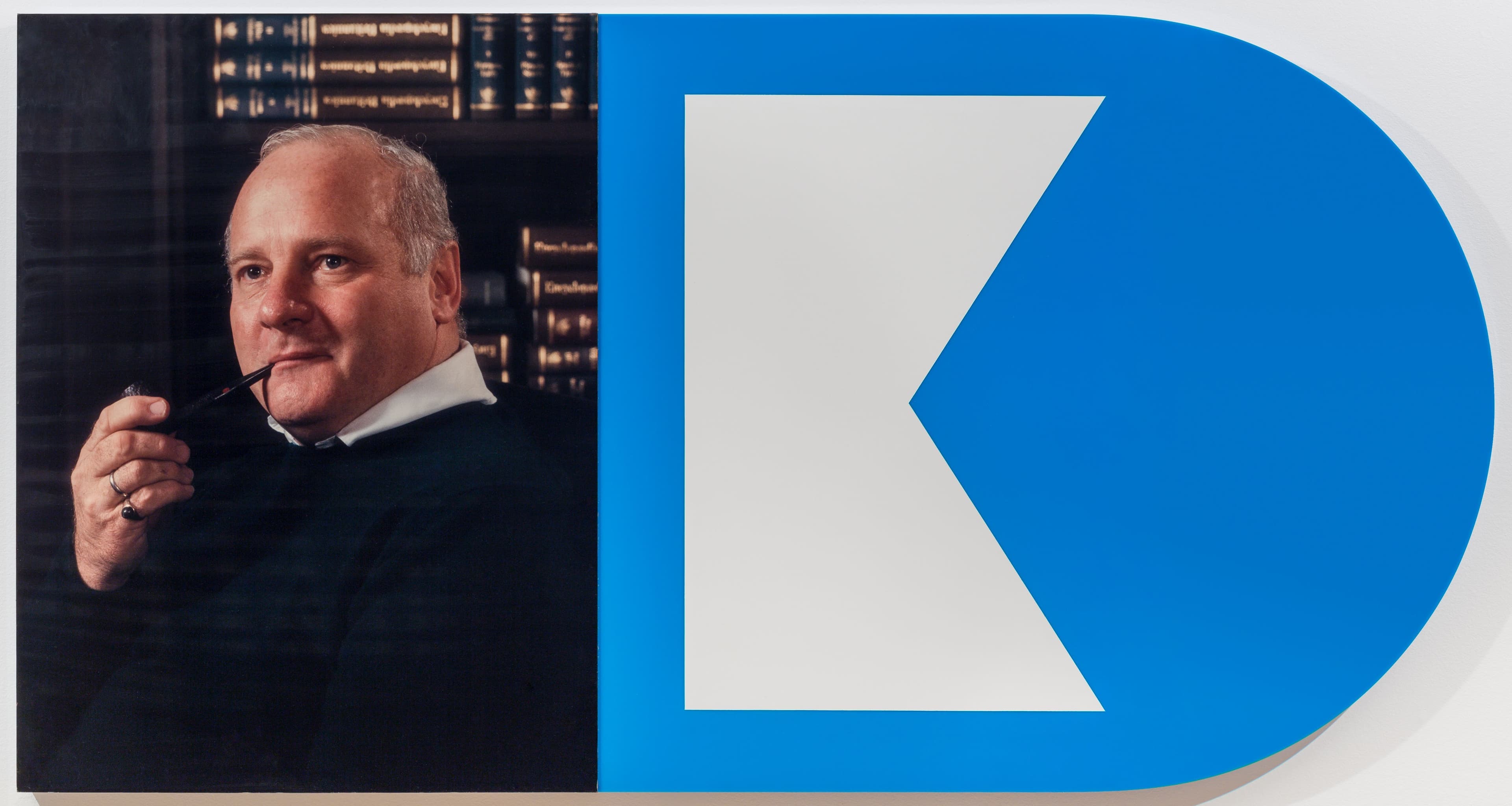 A man with short, light hair and a slight smile holds a pipe in his mouth. He is wearing a black sweater and is positioned against a background of dark wood bookshelves filled with books. To the right, a large blue shape includes a white triangular figure and the letter "K." The overall composition contrasts the portrait with the bold graphic elements.
