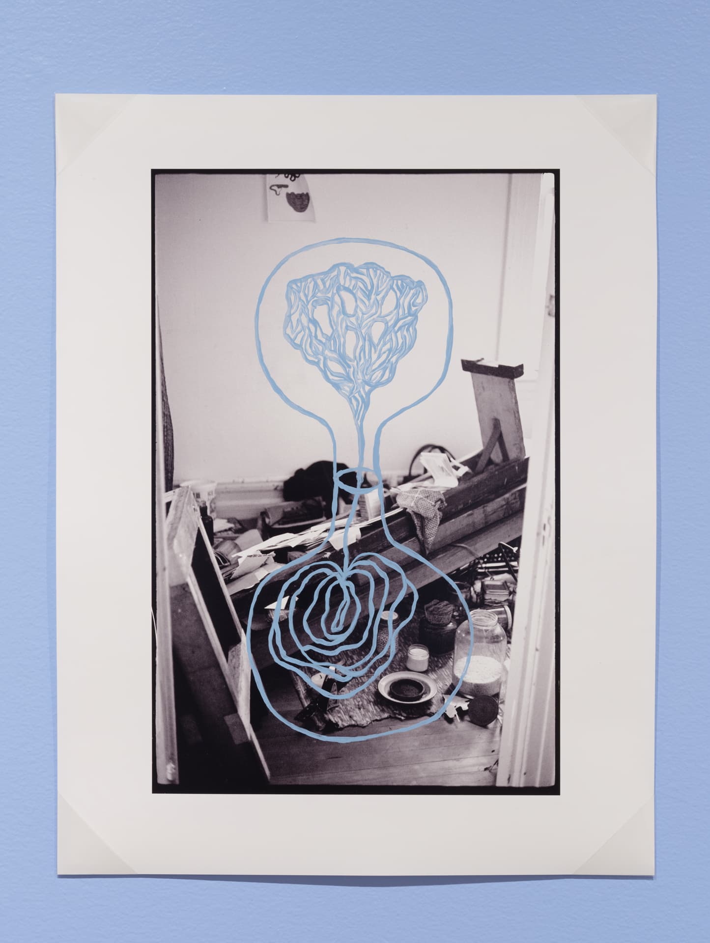 A black-and-white photograph of a cluttered room with scattered objects on the floor. Overlaid on the image is a blue, illustrative design resembling a floral or organic shape, which contrasts with the monochrome scene. The photograph is framed with a white border and is mounted on a light blue wall.