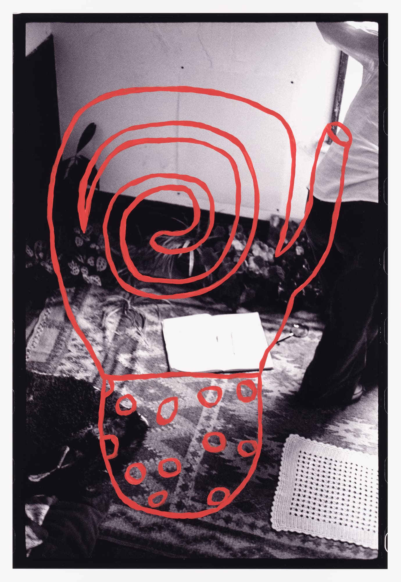 A black and white image featuring a spiral and circular shapes drawn in red overlaid on a patterned rug. In the foreground, there is an open book and a crocheted mat. Parts of a person's legs and feet can be seen in the image. The background includes a wall with a few marks and shadows.
