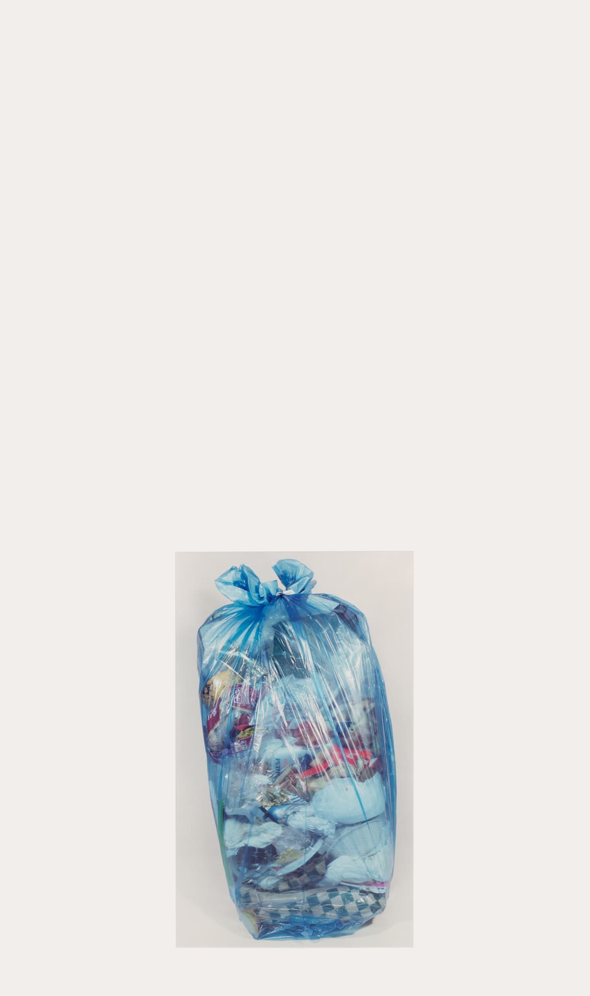 A large, filled garbage bag is shown, made of translucent blue plastic, tied securely at the top. The bag contains various items, some of which are slightly visible through the plastic. The background is a light, neutral color, emphasizing the bag in the center of the image.