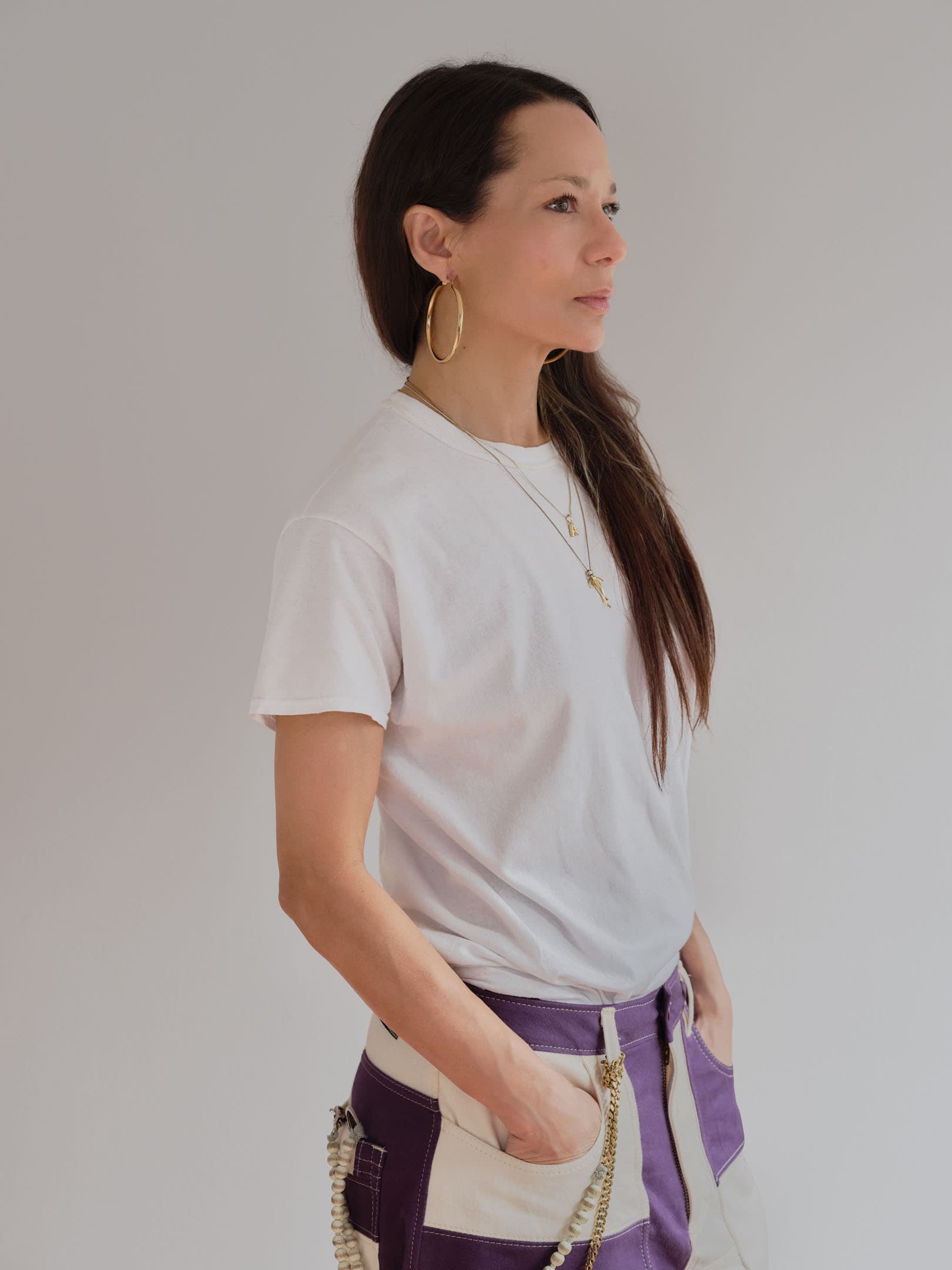 A woman in profile stands against a light gray background. She has long, dark hair and is wearing a plain white t-shirt paired with cream-colored pants that have purple sections. Large gold hoop earrings and several layered necklaces add to her outfit. Her hand rests in the pocket of her pants.