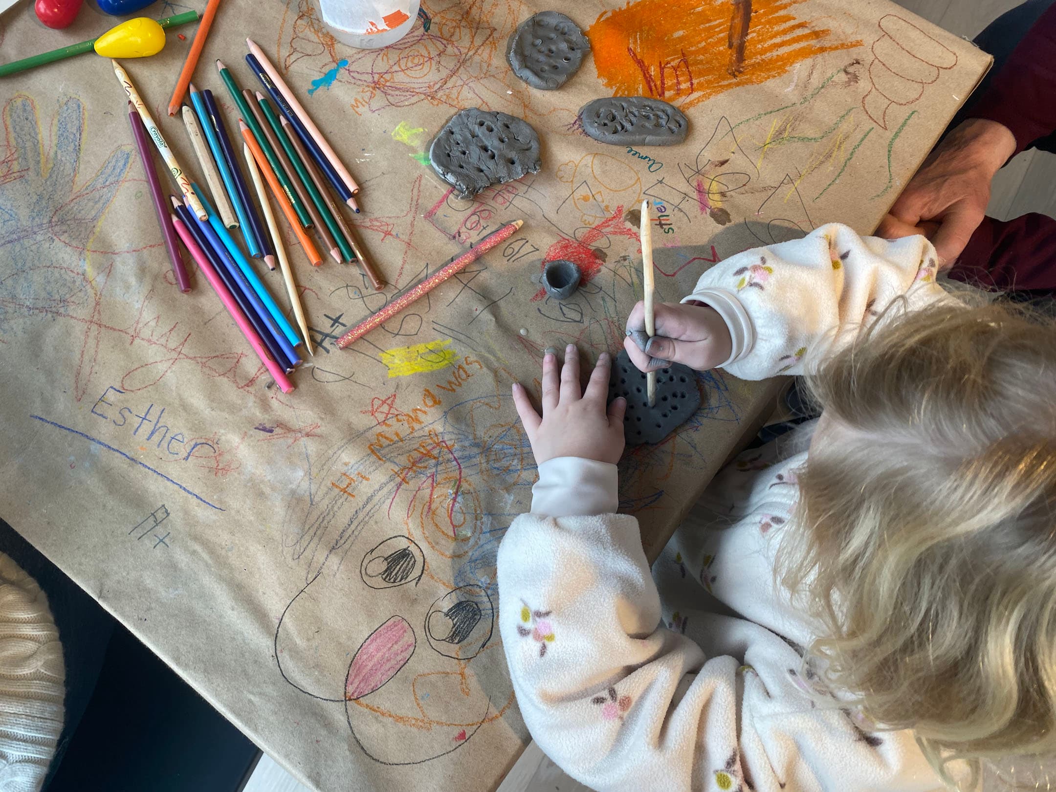 A child with curly hair is sitting at a table covered in brown paper, surrounded by various art supplies including colored pencils, crayons, and small objects. They are using a small stick to work on gray modeling clay, which is shaped into different forms. Colorful scribbles and drawings are visible on the paper, with the name "Esther" written prominently. An adult's hand can be seen assisting or guiding the child from the right side of the image.