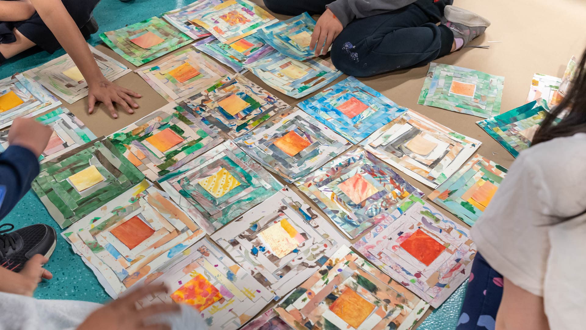 A group of children is sitting on a turquoise floor, surrounded by colorful artwork laid out in a grid pattern. The artwork consists of various painted and collaged pieces with overlapping borders in a range of colors, including orange, green, and blue. Hands are visible as children engage with the art, with some reaching toward the pieces. Brown craft paper is visible beneath the artwork.