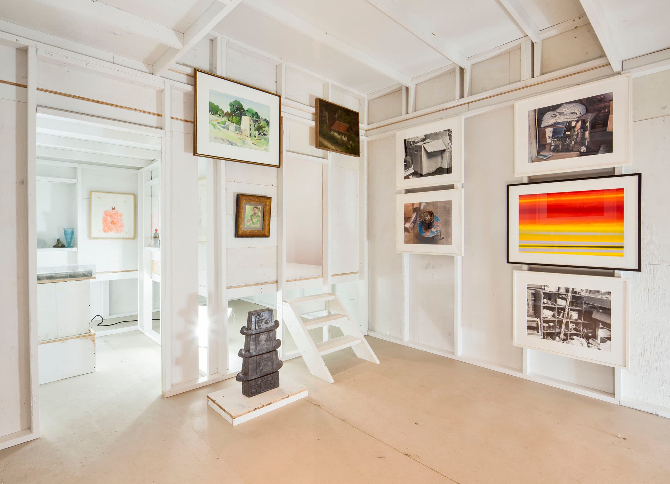 A bright, minimalist art gallery interior with white wooden walls and a light floor. Various framed artworks, including paintings and photographs, are displayed on the walls. A small set of white stairs leads to a raised area, while a dark sculpture is positioned on a pedestal in the foreground. The space is open and well-lit, creating an inviting atmosphere.