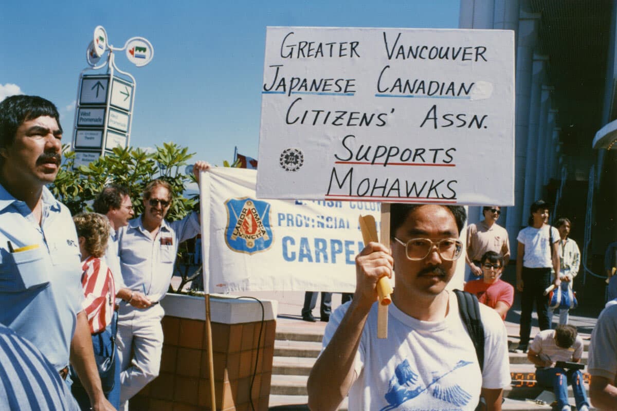 A group of protesters is gathered, with one man in the foreground holding a sign that reads "Greater Vancouver Japanese Canadian Citizens' Assn. Supports Mohawks." The man is wearing glasses and a t-shirt, and there are other individuals behind him holding a banner. The setting appears to be outdoors, with trees and signage visible in the background. Some people are engaged in conversation, while others are looking at their phones or observing the event.