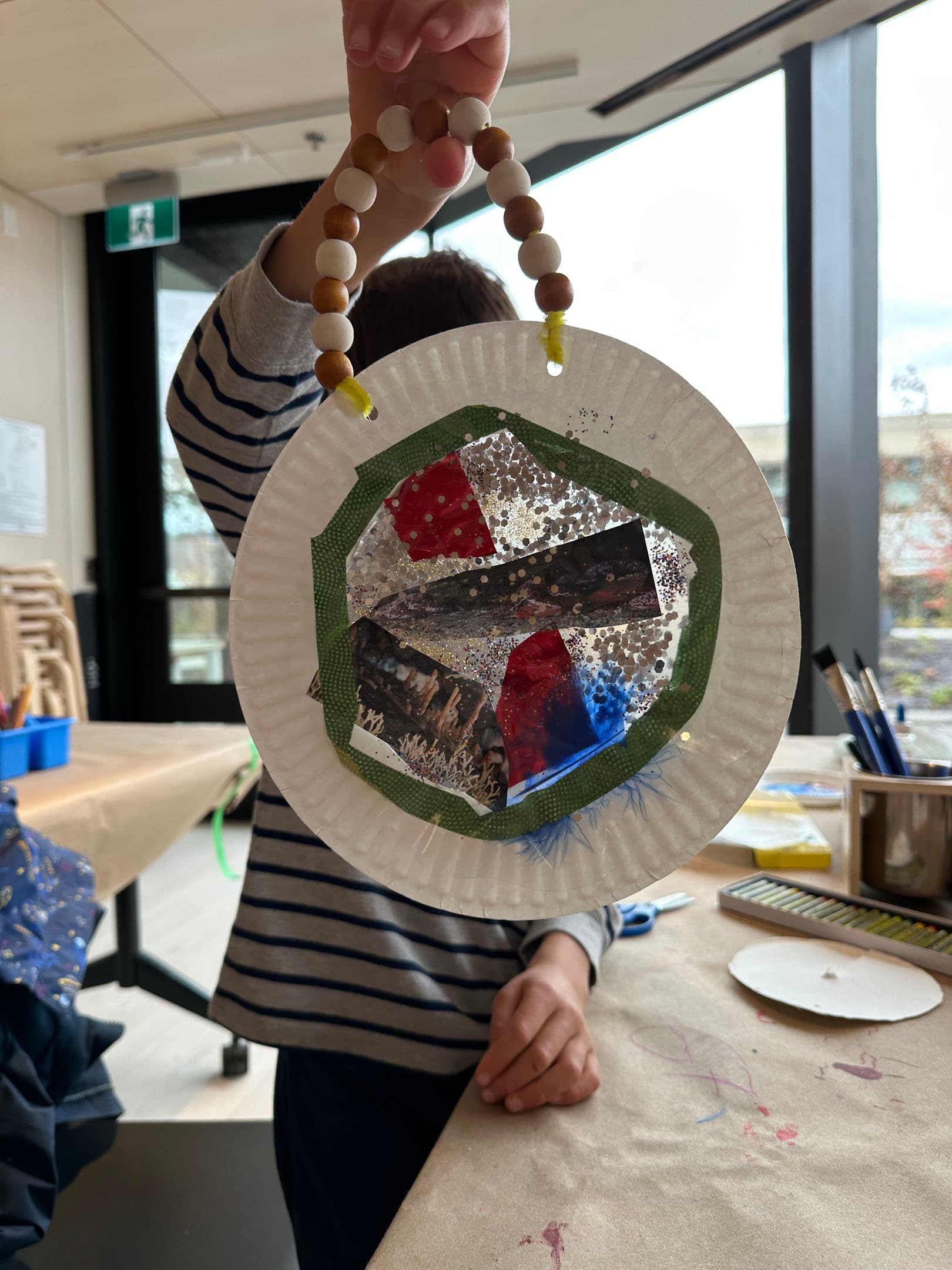 A child is holding a decorated paper plate craft with a beaded handle. The plate features various colorful materials, including shiny and textured elements, predominantly in shades of green, red, blue, and silver. The background shows a classroom setting with tables covered in craft supplies and natural light coming through a large window.