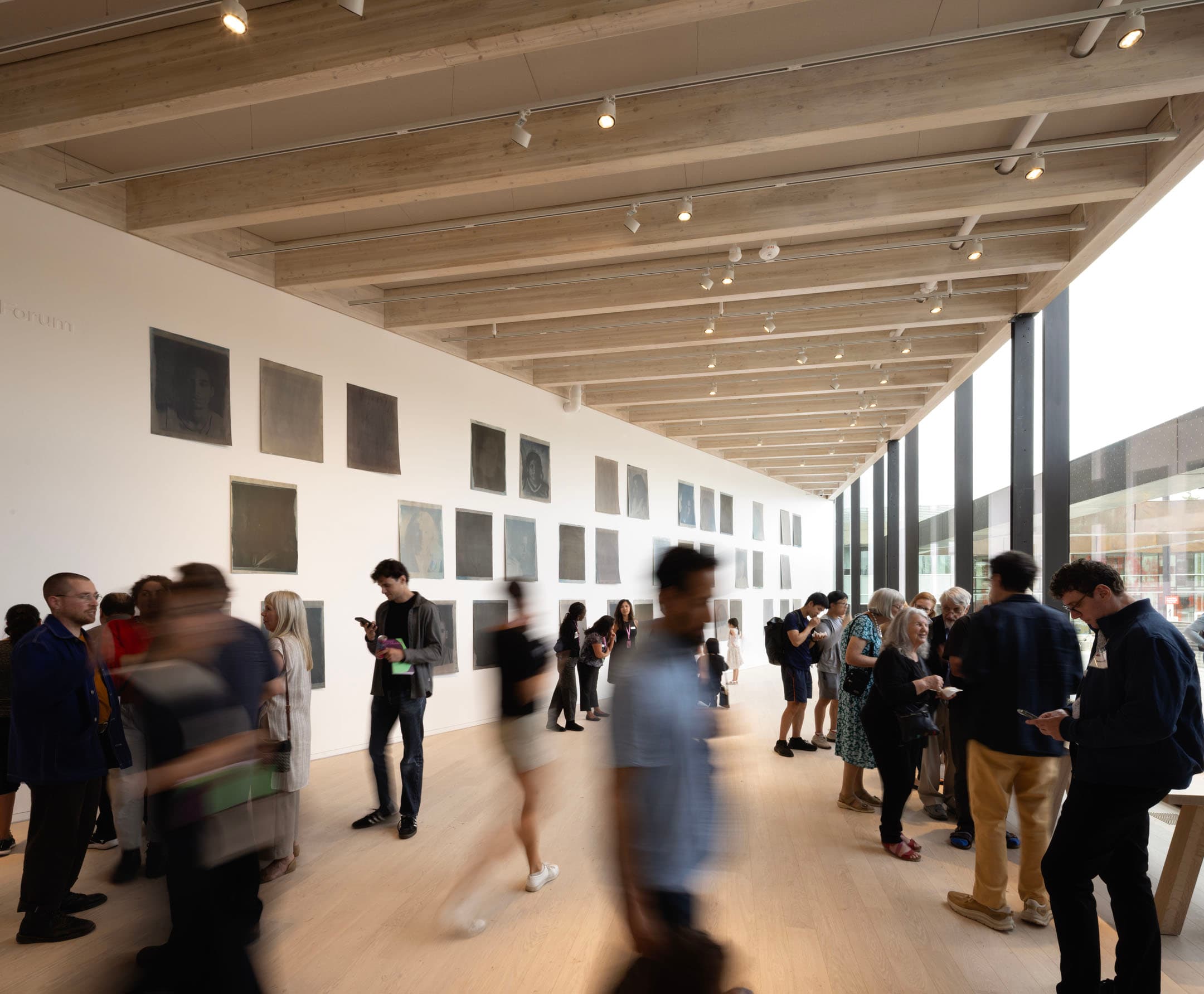 A spacious art gallery features a wooden ceiling and large windows allowing natural light. The walls are adorned with several framed artworks, predominantly in muted tones. A diverse group of people mingles and interacts in the foreground, with some standing still while others are in motion, suggesting a lively atmosphere.