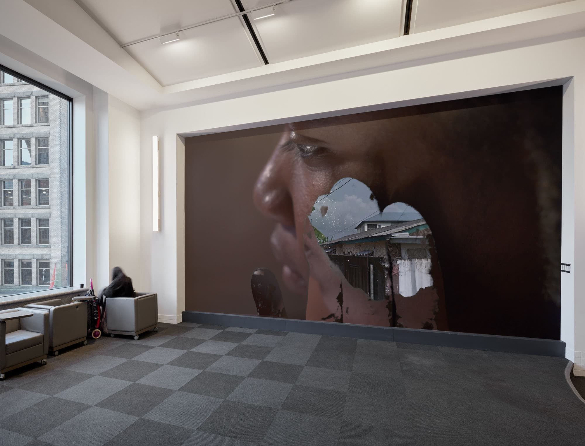 A spacious interior with gray tiled flooring features a large mural on the wall, depicting a close-up of a person's profile alongside an image of a building in the background. Natural light enters through a window on the left, where a person sits in a gray armchair. The overall atmosphere is modern and minimalist, with neutral color tones.