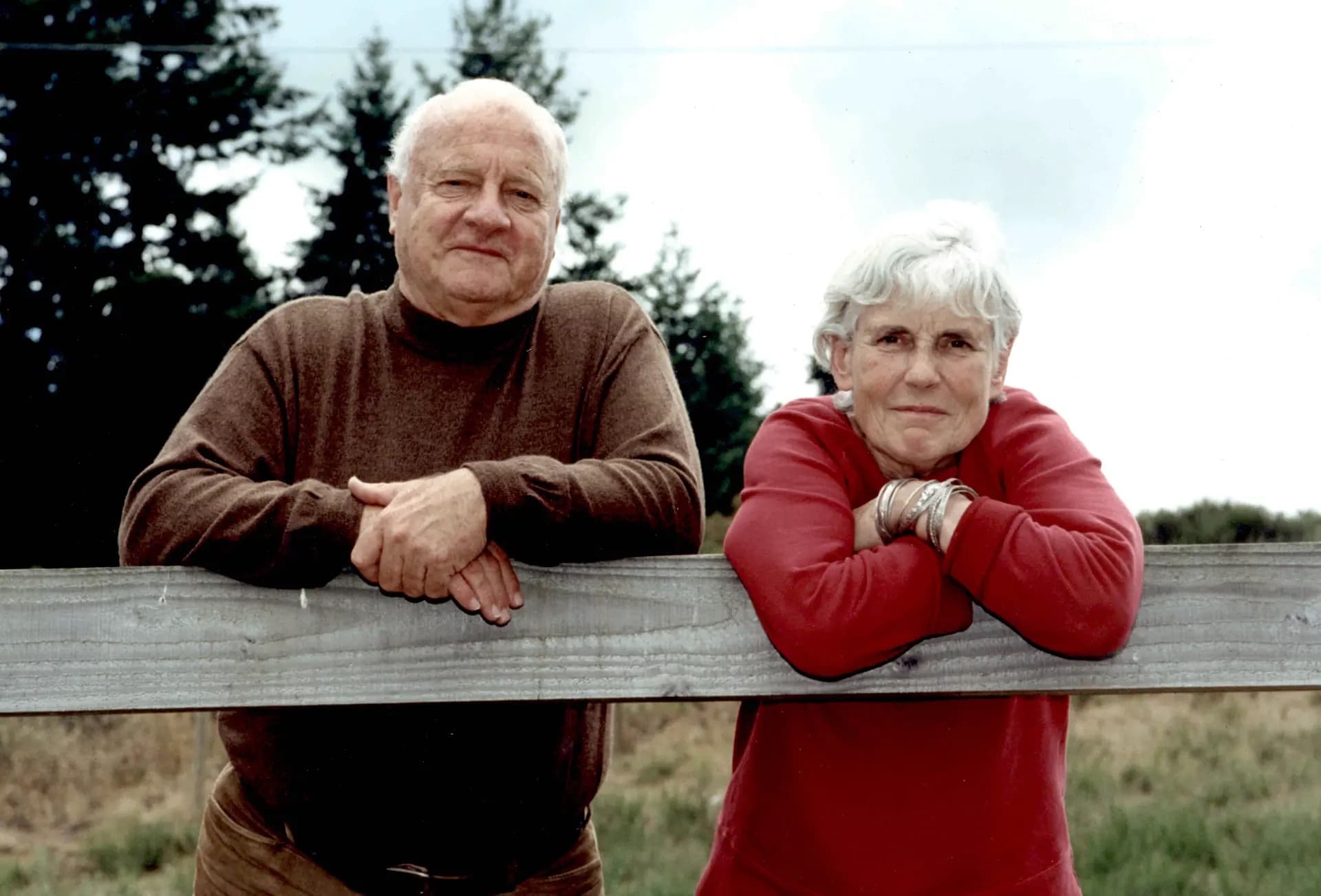 An elderly man and woman stand together by a wooden fence, with the man wearing a brown turtleneck sweater and the woman wearing a red sweatshirt. They both lean on the fence, smiling gently. In the background, there are trees and a cloudy sky. The scene conveys a warm, intimate moment between them.