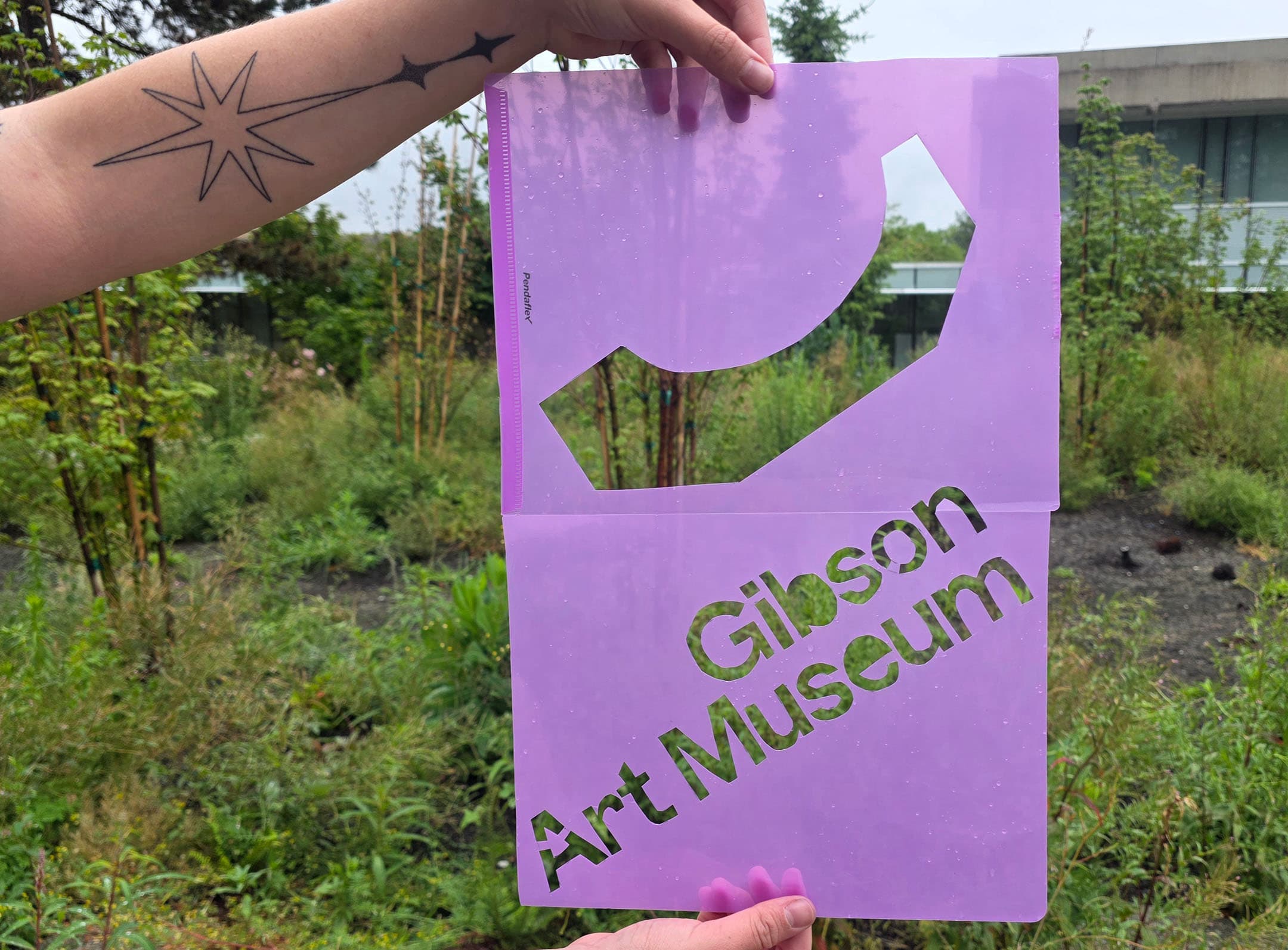 A hand holds a translucent purple sheet with cut-out shapes and the text "Gibson Art Museum" displayed prominently. In the background, greenery and plants are visible, indicating an outdoor setting. The hand also features a tattoo of a star.