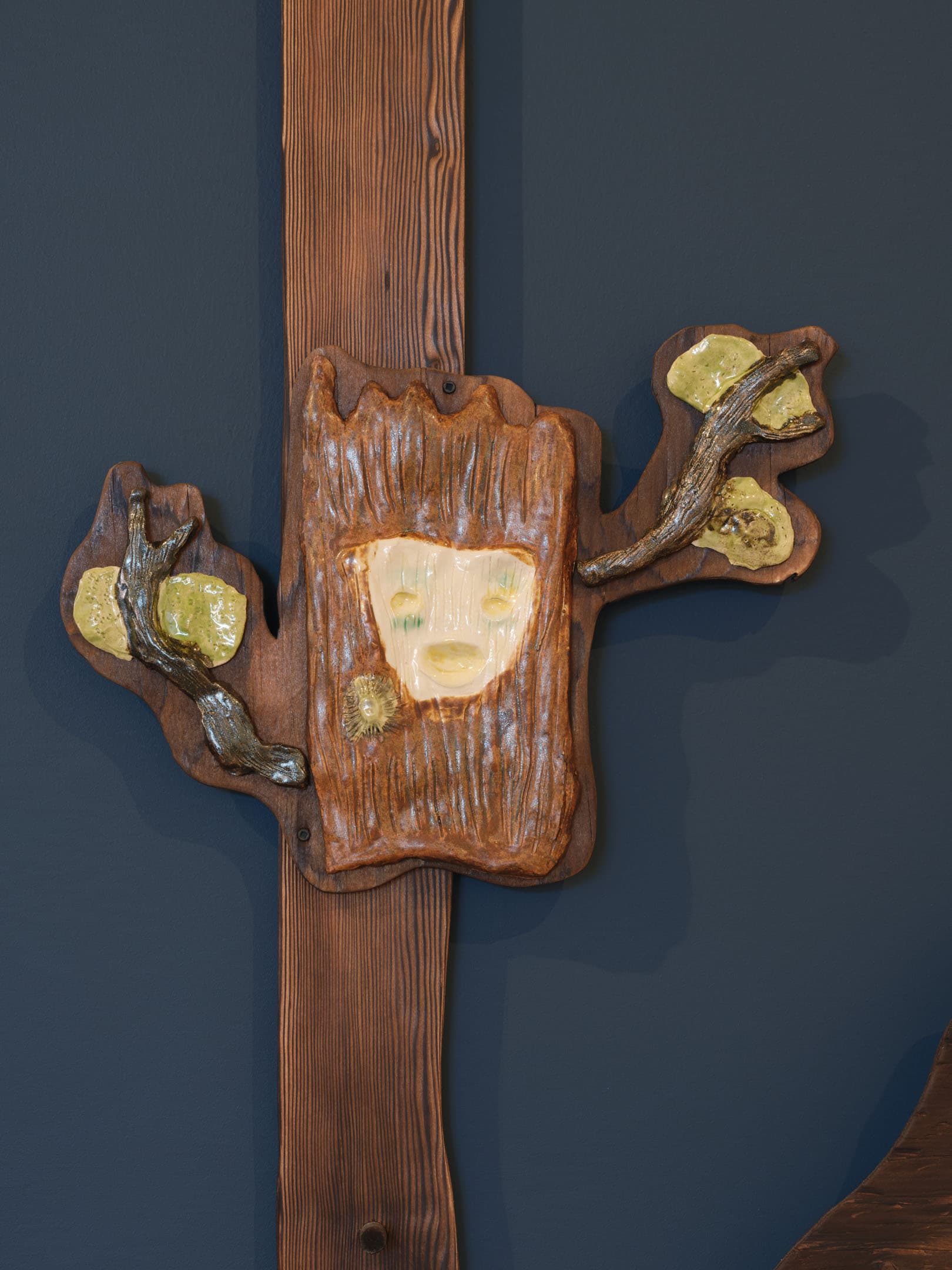 A decorative wall piece resembling a tree stump with a face carved into it. The piece features textured brown wood and is adorned with green and yellow accents resembling leaves. It is mounted on a dark blue wall, with a vertical wooden beam beside it.