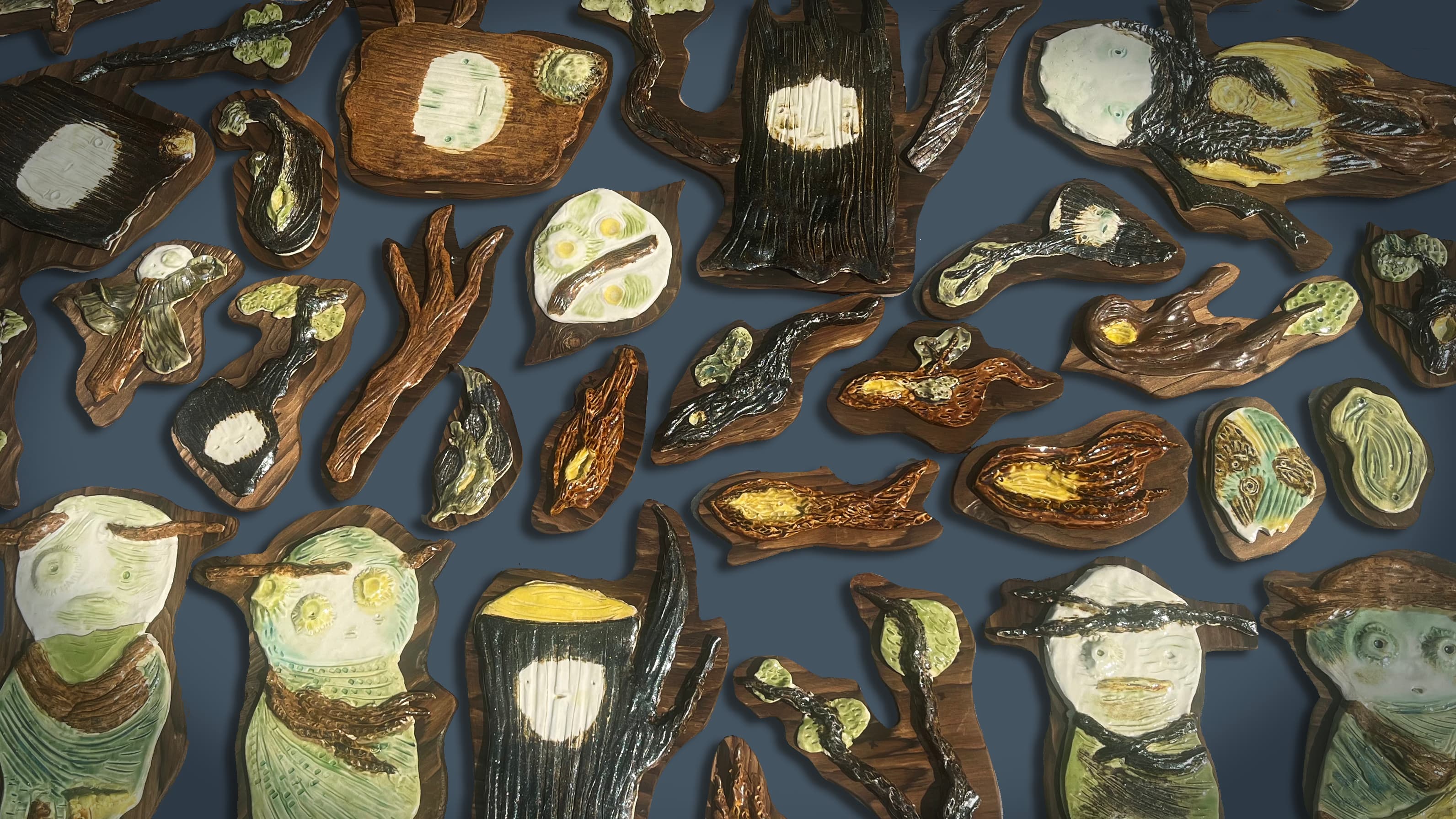 A collection of intricately designed ceramic pieces resembling abstract figures, trees, and organic shapes, arranged flat on a dark background. The pieces showcase a variety of textures and colors, including browns, greens, yellows, and whites, emphasizing a natural and artistic aesthetic.