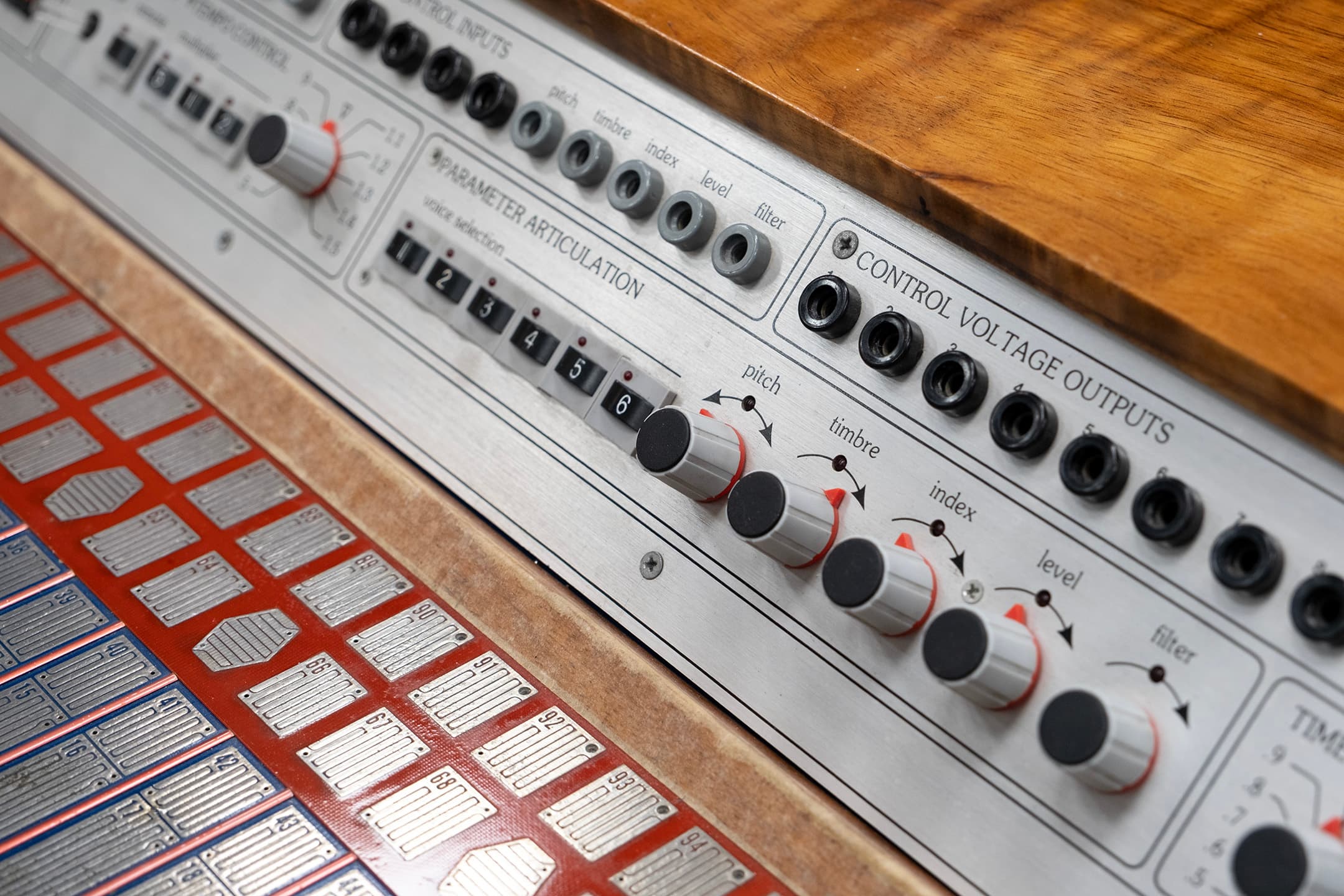Close-up of a synthesizer interface featuring various control knobs and input/output jacks. The panel includes labeled sections for "Control Inputs," "Control Voltage Outputs," and "Parameter Articulation," with adjustable knobs for pitch, timbre, index, level, and filter settings. Below, there are metal plates with numbered labels arranged in rows. The surface is made of wood, providing a warm contrast to the metallic components.