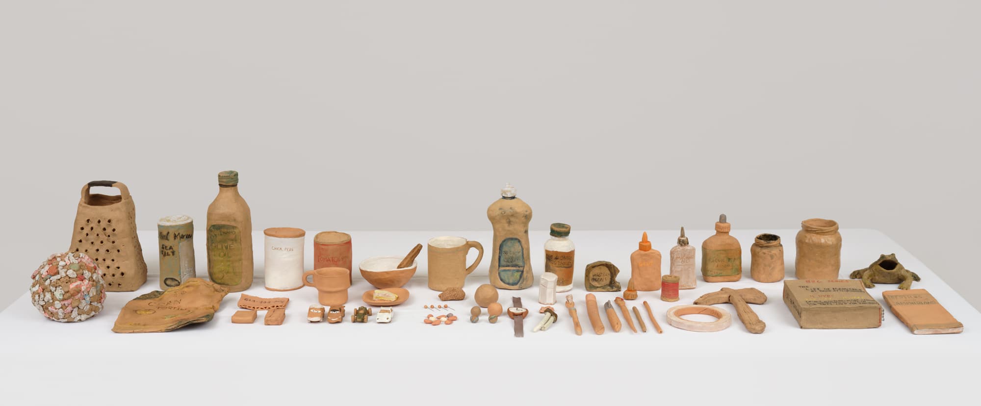 A collection of various pottery items is displayed on a white table against a light gray background. The items include a grater, several bottles labeled for different substances (such as olive oil and sea salt), jars, a bowl with a spoon, plates, and small figurines. The pieces vary in shape and size, with some featuring detailed labels and embellishments. The overall arrangement is organized and showcases the artistic craftsmanship of the ceramic objects.