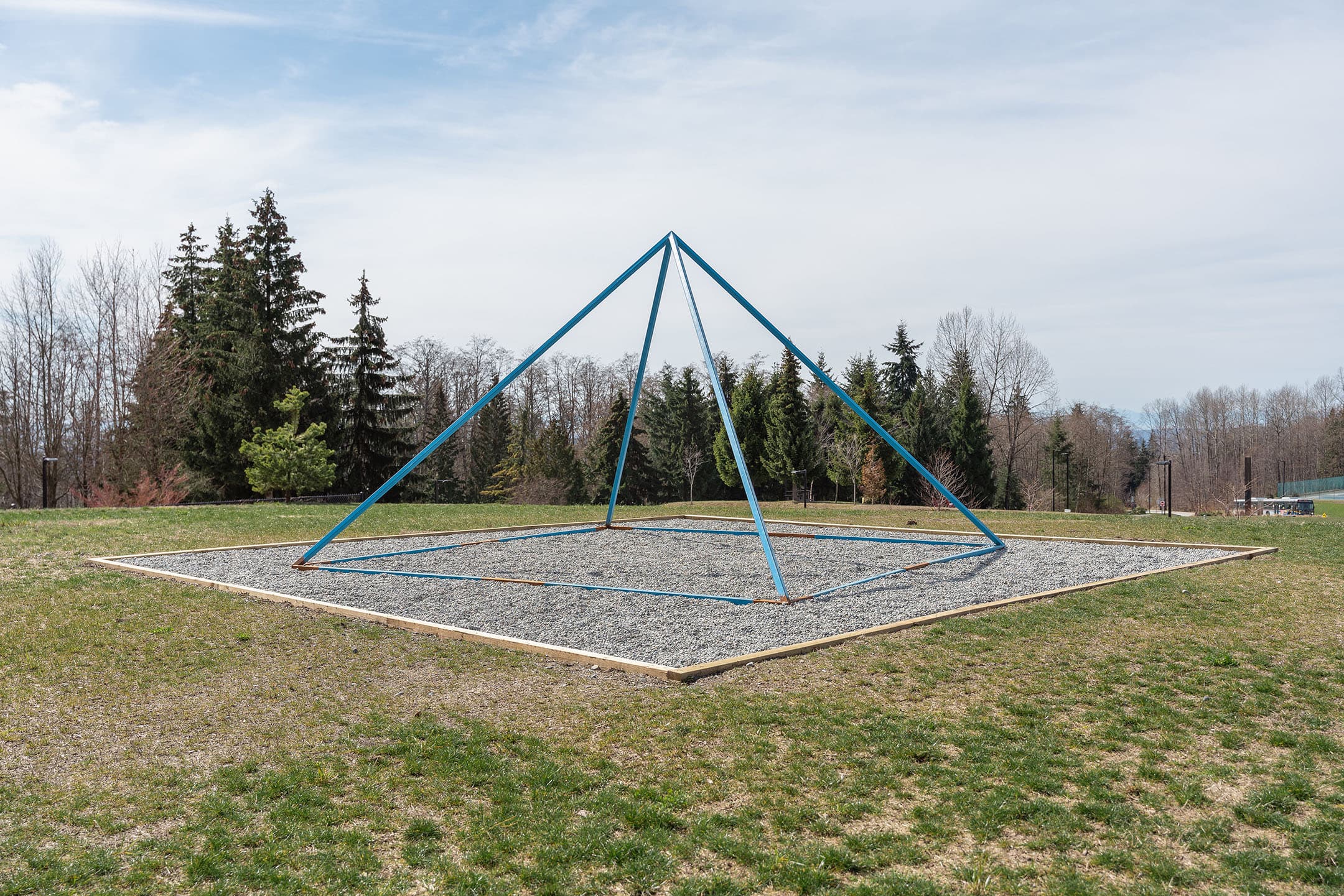 A large blue pyramid-shaped climbing structure sits on a gravel-lined square in a grassy park. The background features tall green trees and a blue sky with some clouds. The area is relatively empty, emphasizing the unique structure.