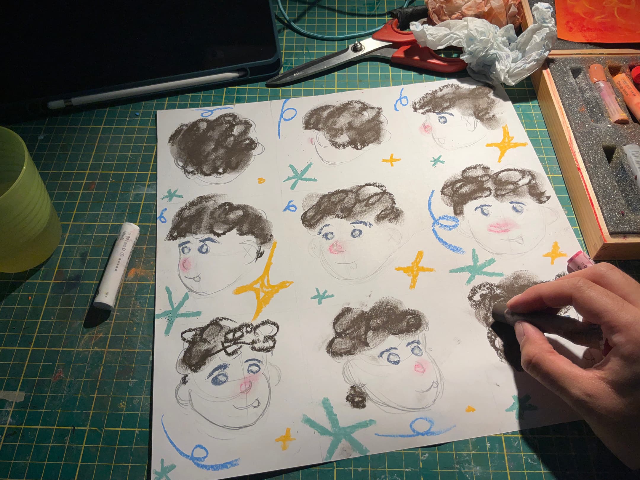A tabletop scene featuring a partially completed drawing on white paper. The drawing consists of multiple cartoon-style faces depicted in soft colours. Surrounding the faces are abstract shapes in yellow, green, and blue. A hand is shown using a black chalk-like medium to add details to one of the faces. Nearby, there are art supplies, including a yellow cup, a white marker, and a wooden box containing pastels. The tabletop has a green cutting mat underneath.