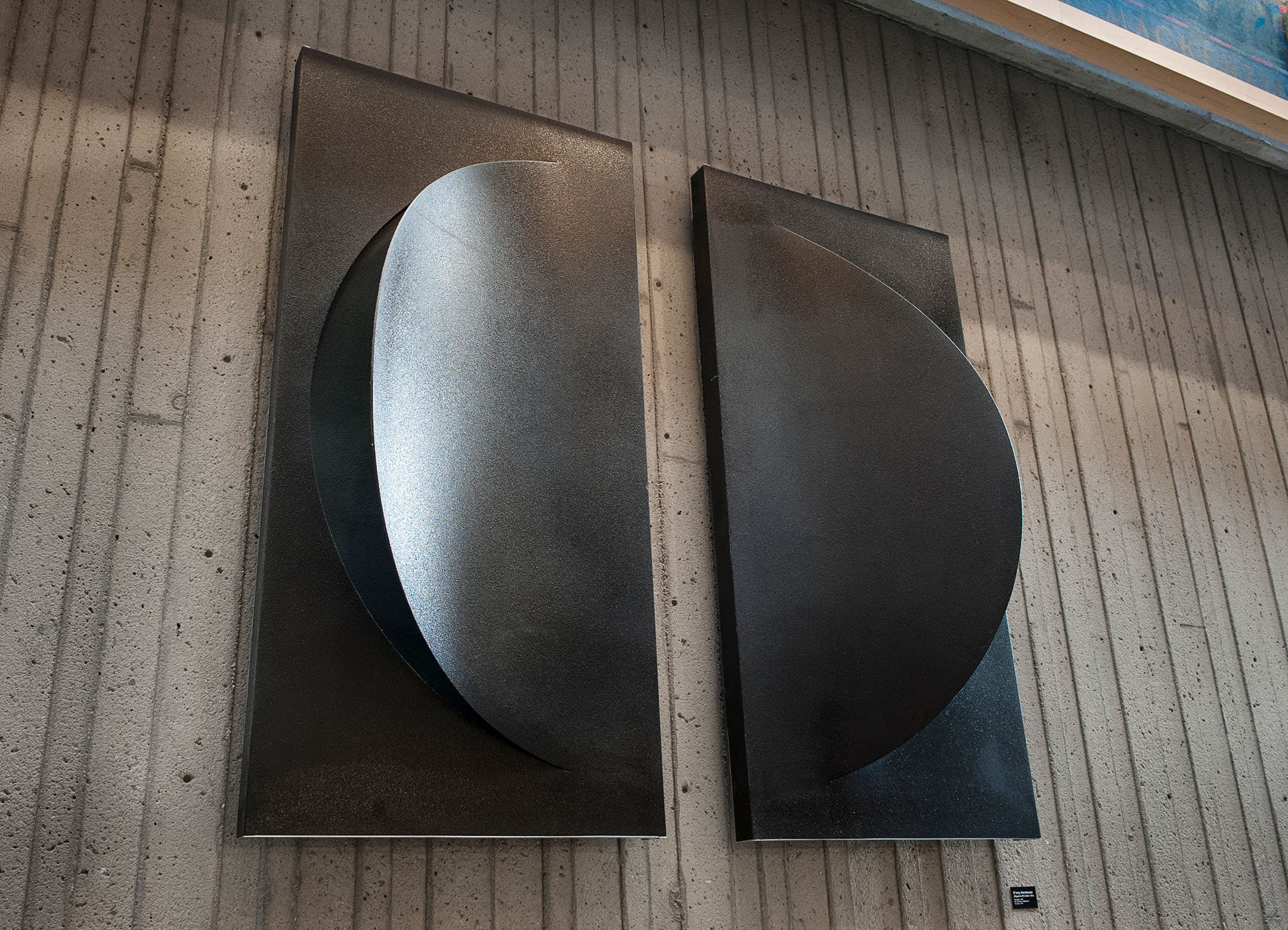 Two large, abstract black wall panels are mounted on a textured concrete wall. Each panel features a smooth, curved shape that reflects light subtly, creating a sense of depth. The panels are positioned closely together, with one featuring a more pronounced curve than the other. A small label can be seen at the bottom right of the left panel.