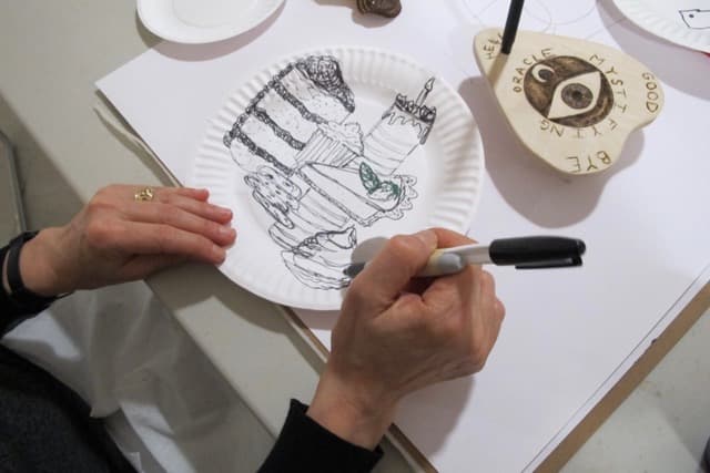 A person's hand is seen drawing on a paper plate with a black marker, creating an illustration of a multi-layered cake adorned with decorative elements. There are additional plates and objects in the background, including an item with text that reads "ORACLE" and a small figurine. The person's wrist is adorned with a bracelet, and they are wearing a black long-sleeved shirt.