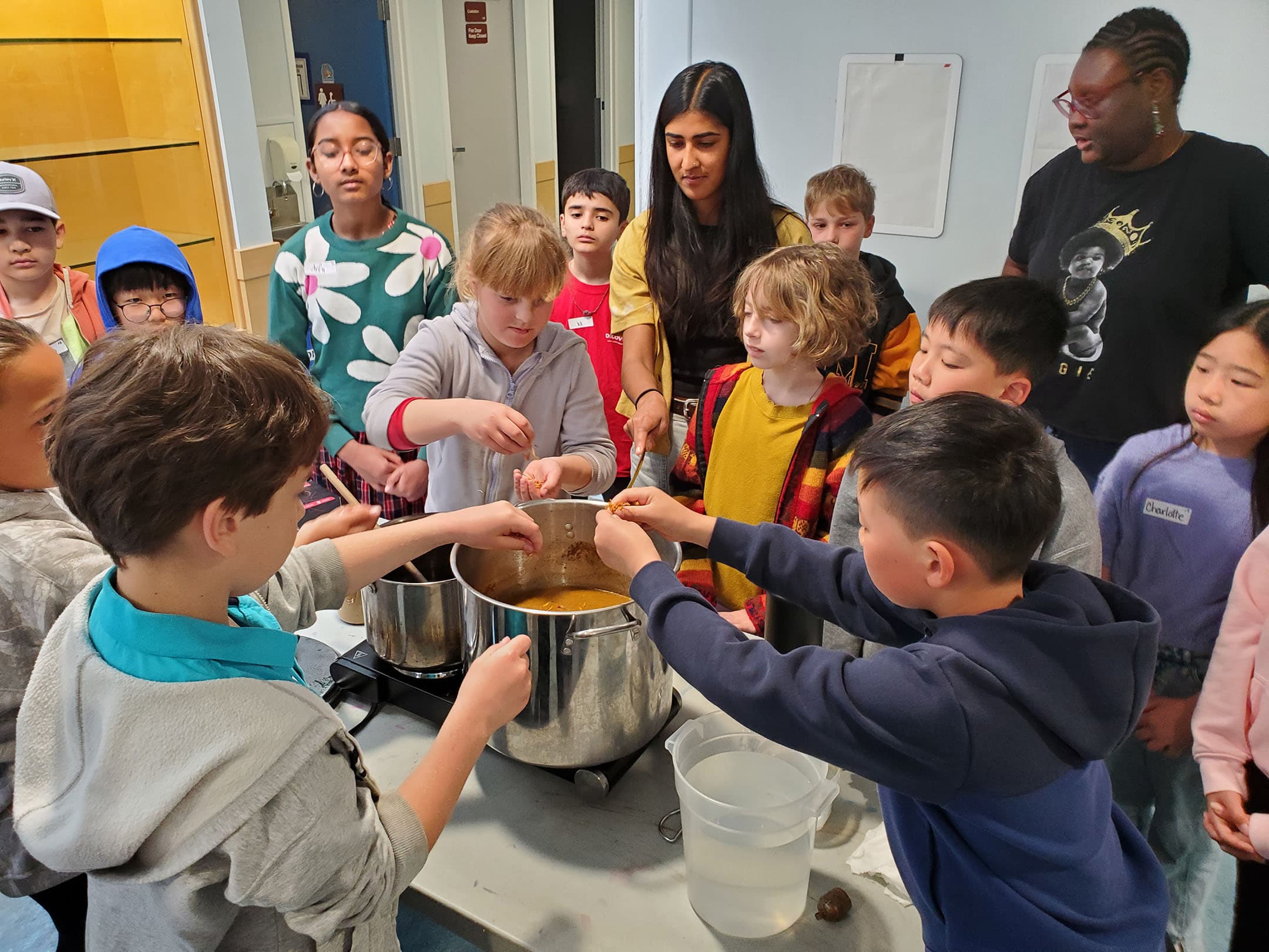 A group of children, along with two adults, are gathered around a table engaged in a cooking activity. Several kids are reaching into a large pot with a brown mixture, while one child also holds a measuring cup filled with water. The background features a colorful wall and a poster board. The children exhibit varying expressions of curiosity and focus as they participate in the activity.