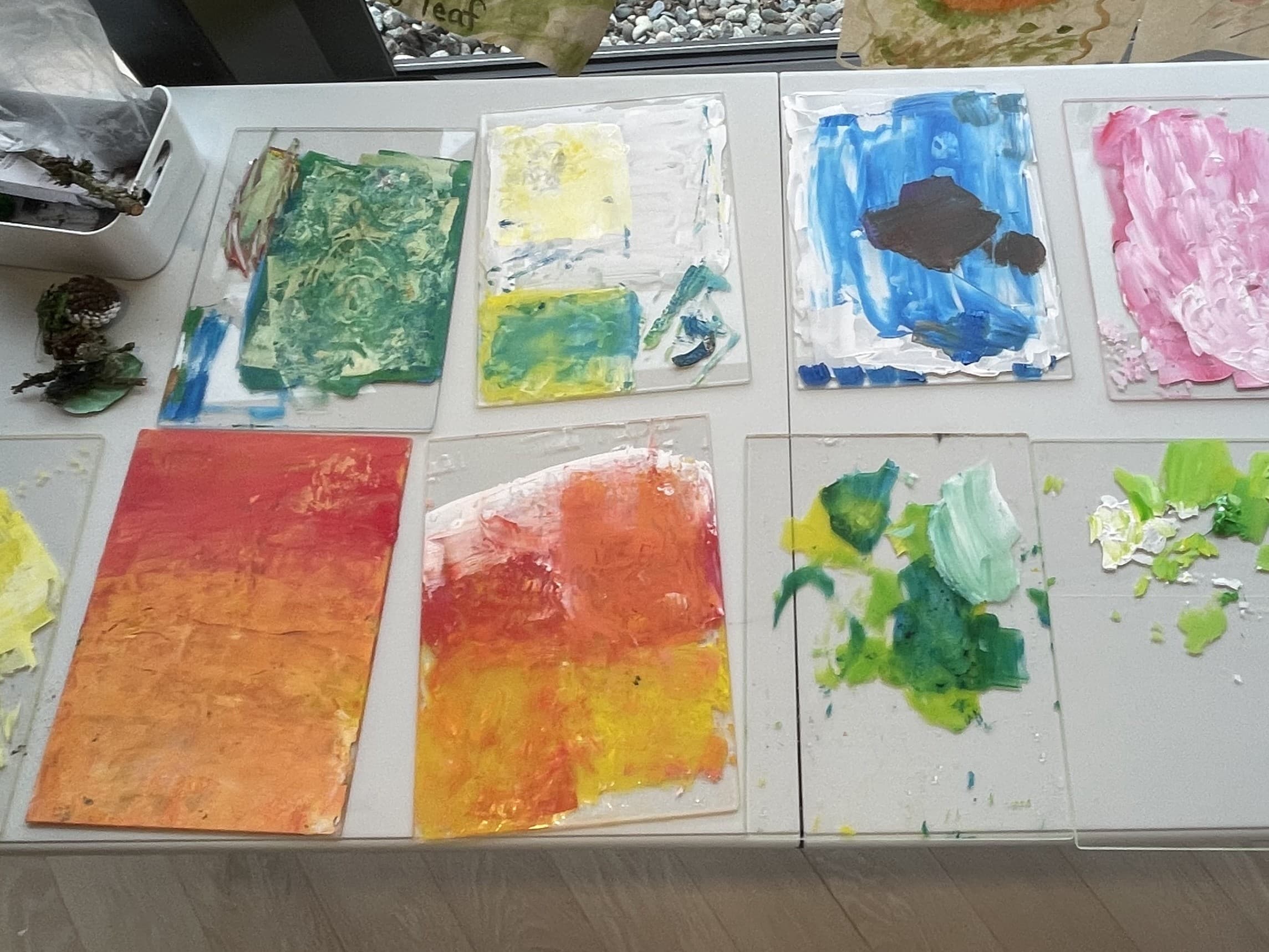 A collection of colorful, textured paintings displayed on a table. The artworks feature various shades, including green, blue, orange, pink, and yellow, with some having visible brush strokes. The setting has a bright, airy atmosphere with a window showing a stone landscape outside.