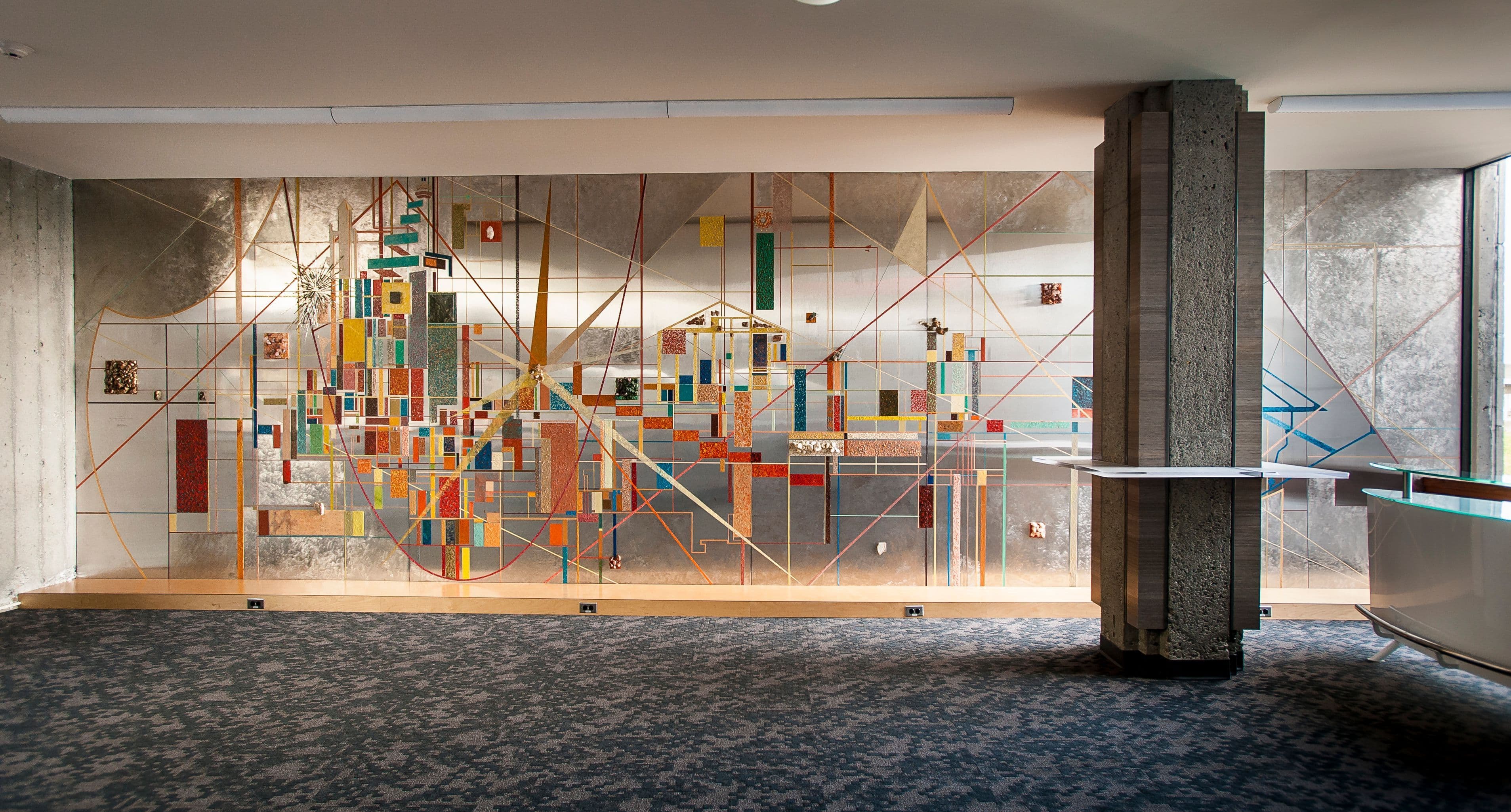 A large, colorful abstract mural is displayed on a wall, featuring geometric shapes and patterns in various colors, including red, yellow, blue, and gold. The mural is contrasted against a background of silver and gray tones. In the foreground, there is a textured gray carpet, along with a concrete pillar and a white table-like structure. Natural light streams in from a window, illuminating the scene.