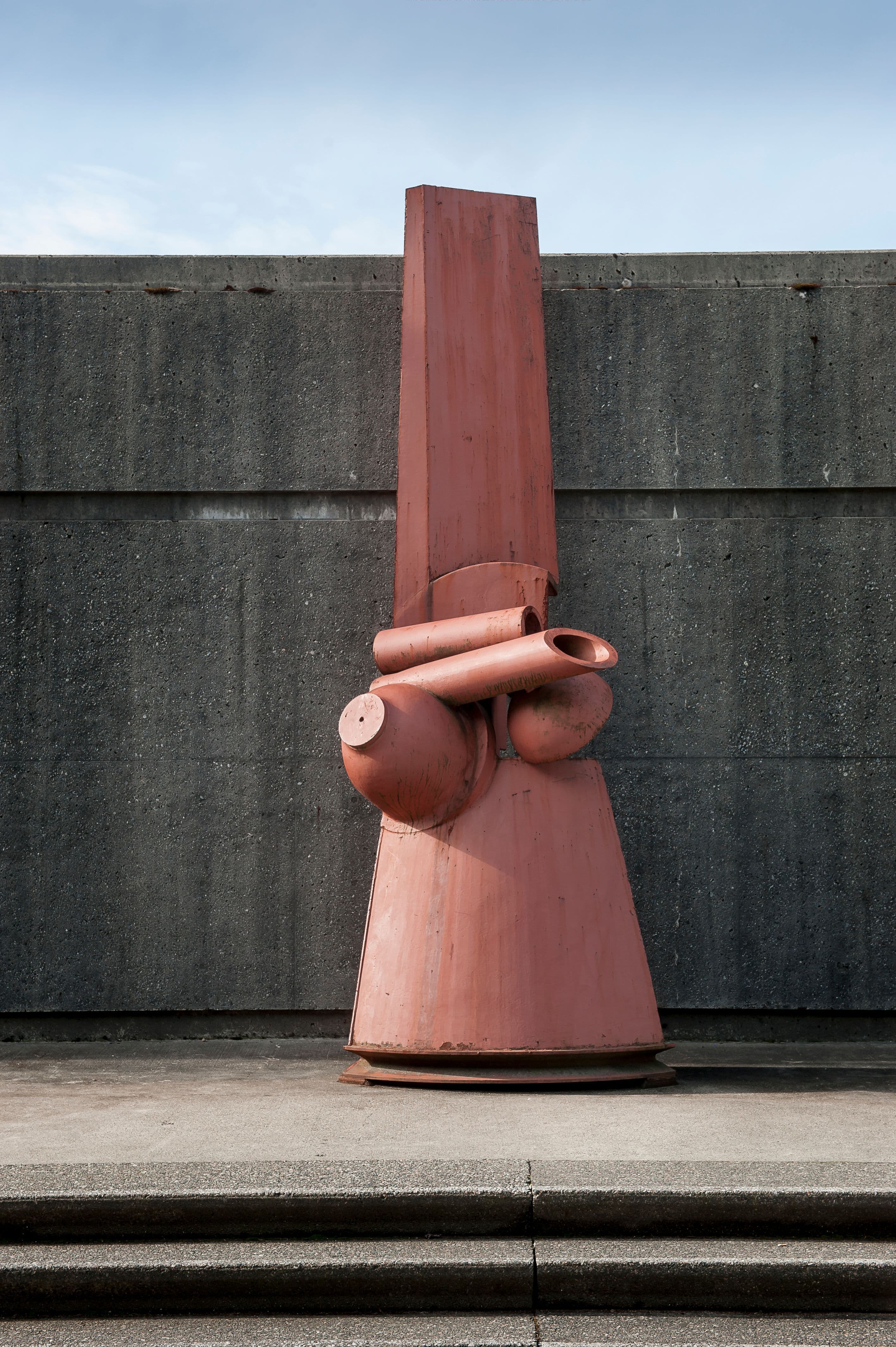 A large, abstract sculpture in an orange-pink hue stands against a textured gray concrete wall. The sculpture features various rounded and cylindrical shapes, with a prominent vertical element at the top and wider forms at the base. The ground below the sculpture consists of concrete steps leading up to it. The sky above is predominantly blue with a few clouds.