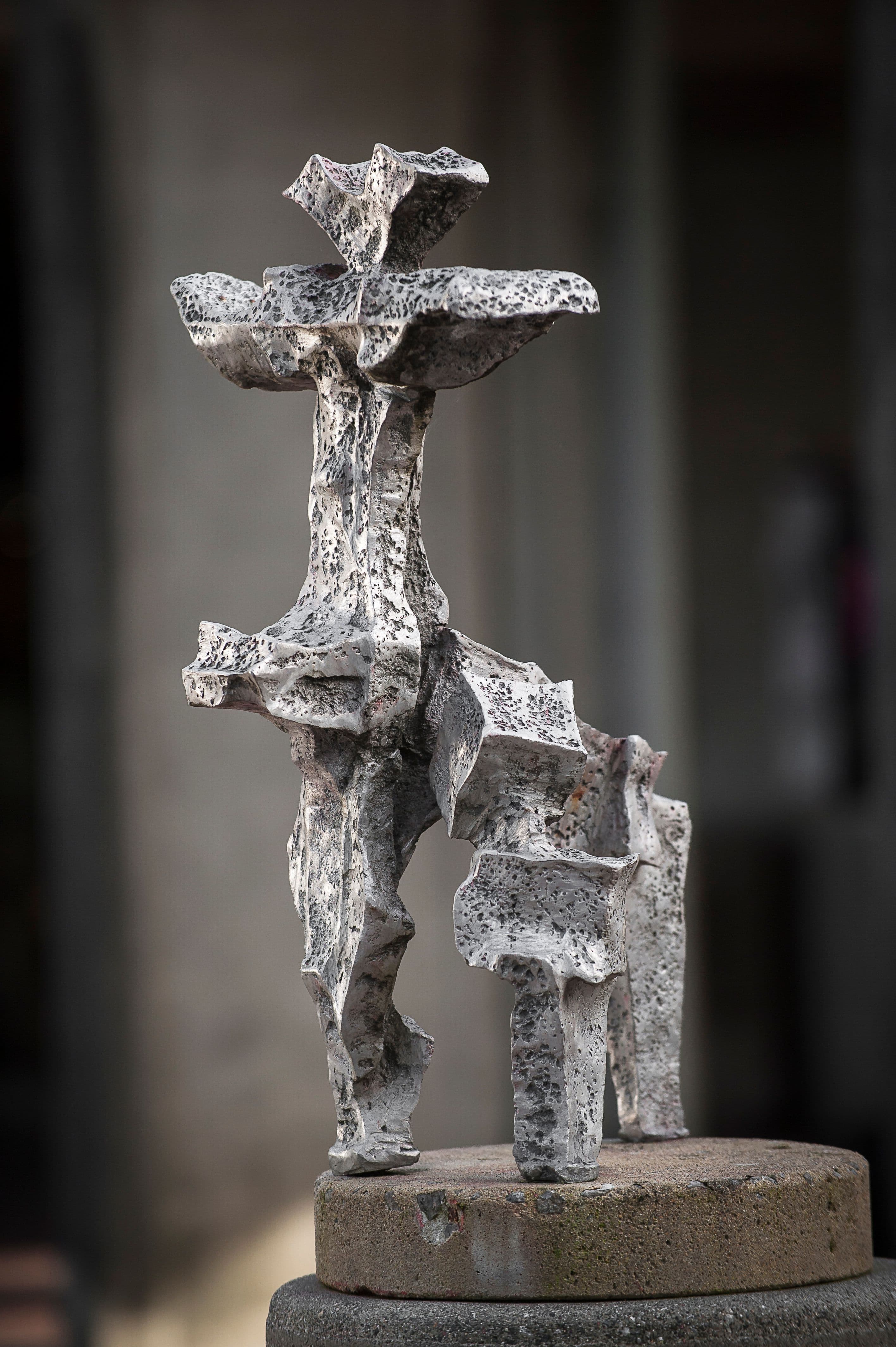 A textured, abstract sculpture stands on a round stone base. The sculpture features irregular, jagged forms with a mix of gray and white colors, creating a dynamic, three-dimensional appearance. It has a layered top that resembles ruffled edges, with figures extending in various directions. The background is softly blurred, emphasizing the artwork.