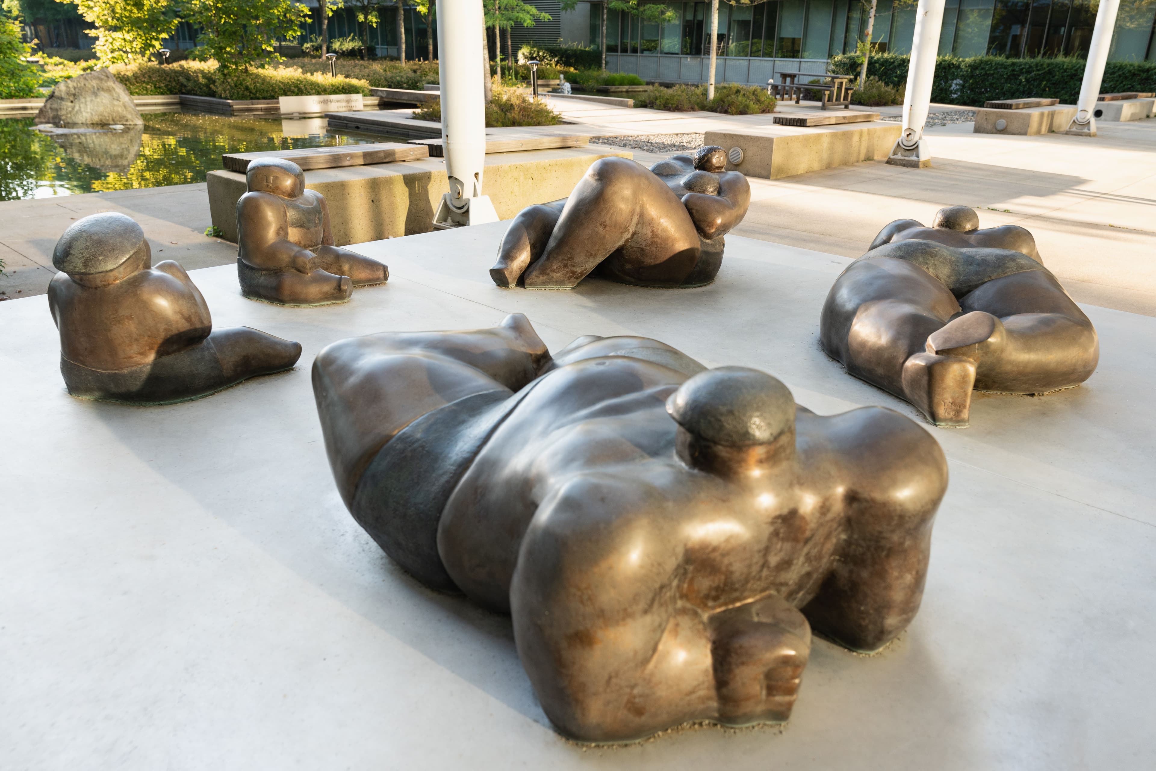 A group of five bronze sculptures is arranged in a modern outdoor setting. Each sculpture depicts abstract, rounded human forms in various reclining poses, resting on a smooth, light-colored concrete surface. The background features greenery, including trees and a body of water, with a building visible in the distance.
