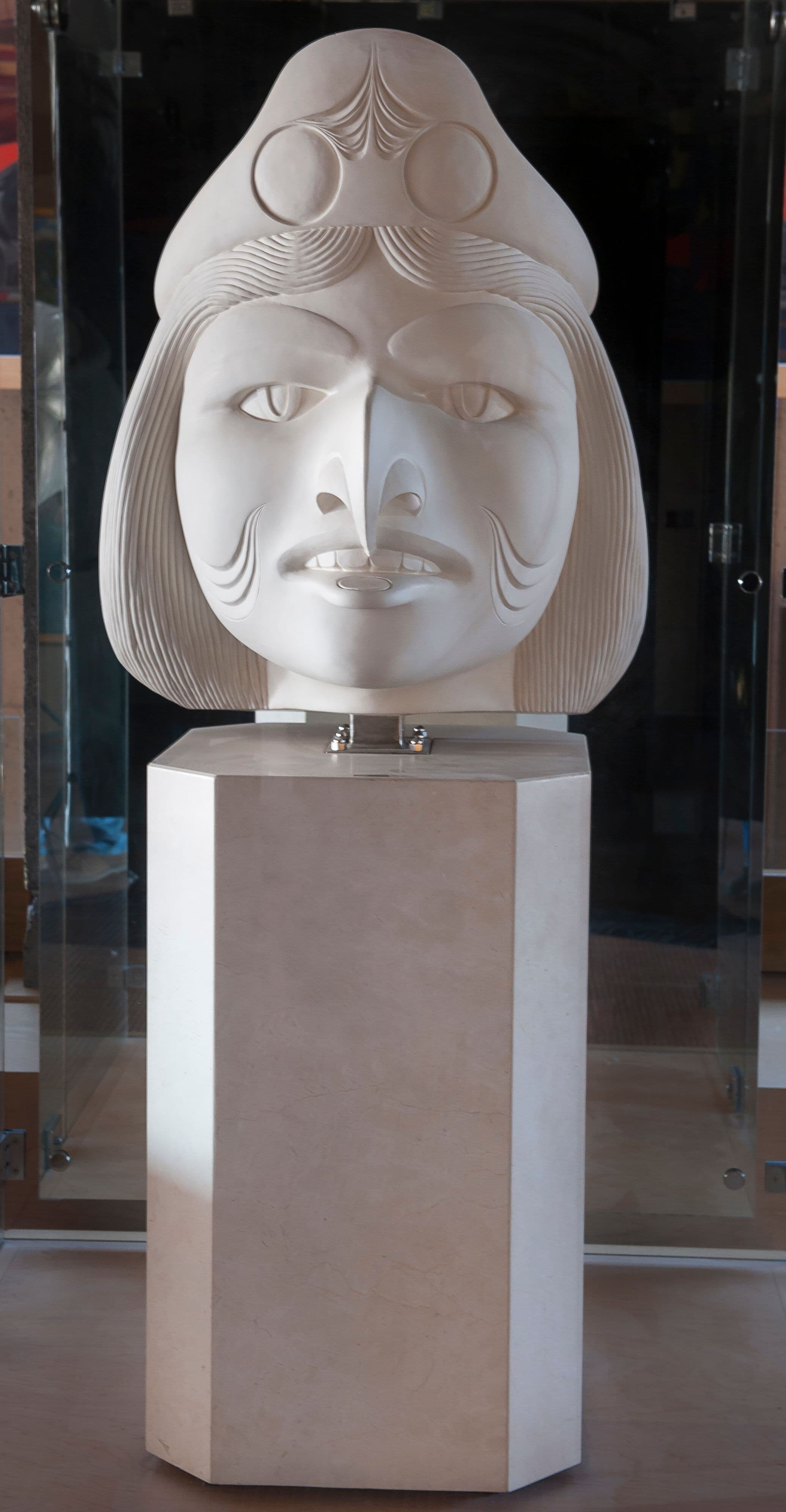 A large, stylized sculpture of a face, made of white stone, is displayed on a rectangular pedestal. The face features prominent eyes, a defined nose, and a mouth with a slight smile. The figure is adorned with a headdress that includes circular designs. The background has glass elements that suggest an indoor exhibition space.